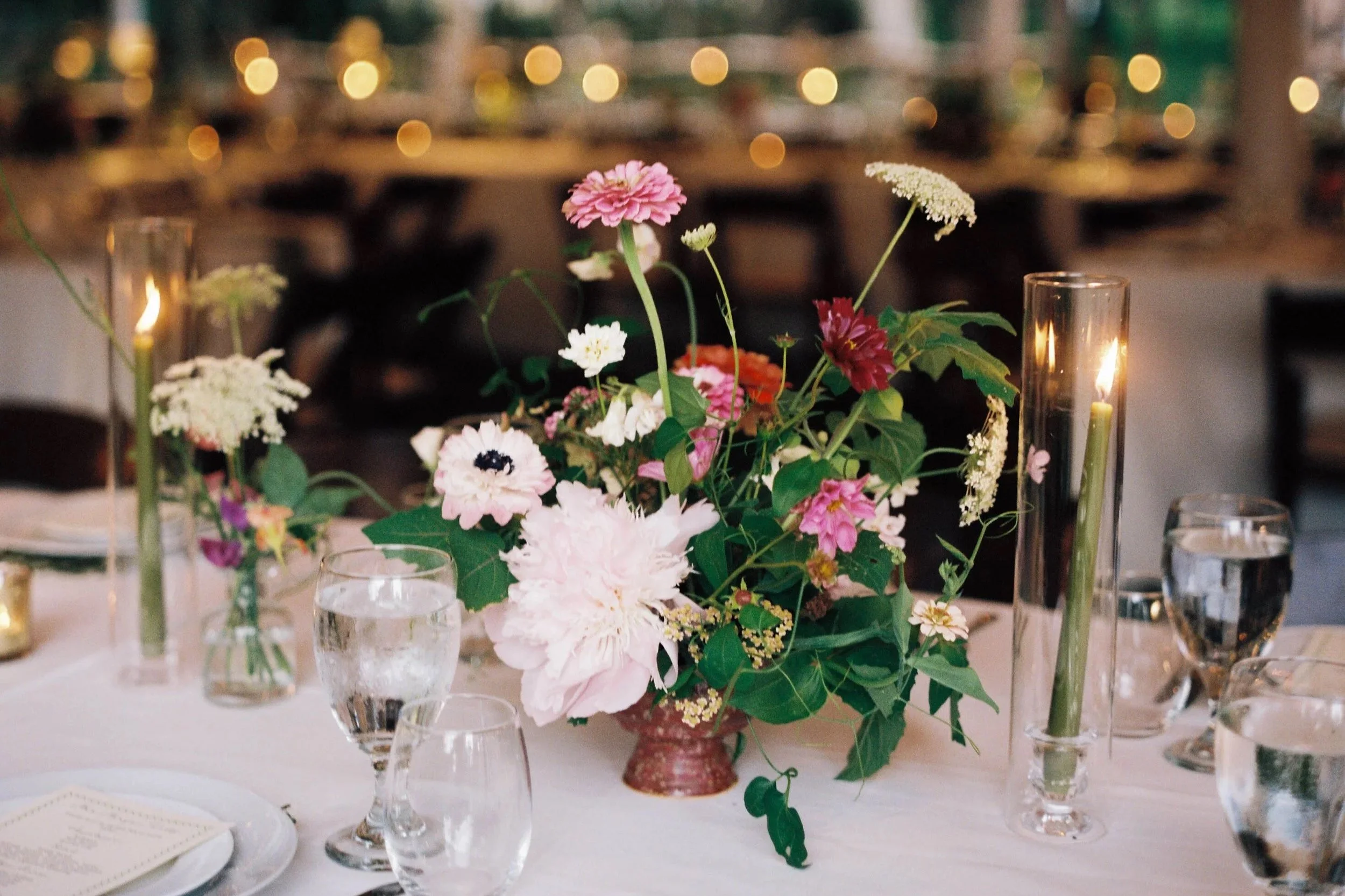 A long wooden banquet table decorated with white flowers, candles, and gold accents in a rustic wooden venue with large windows and white chairs.