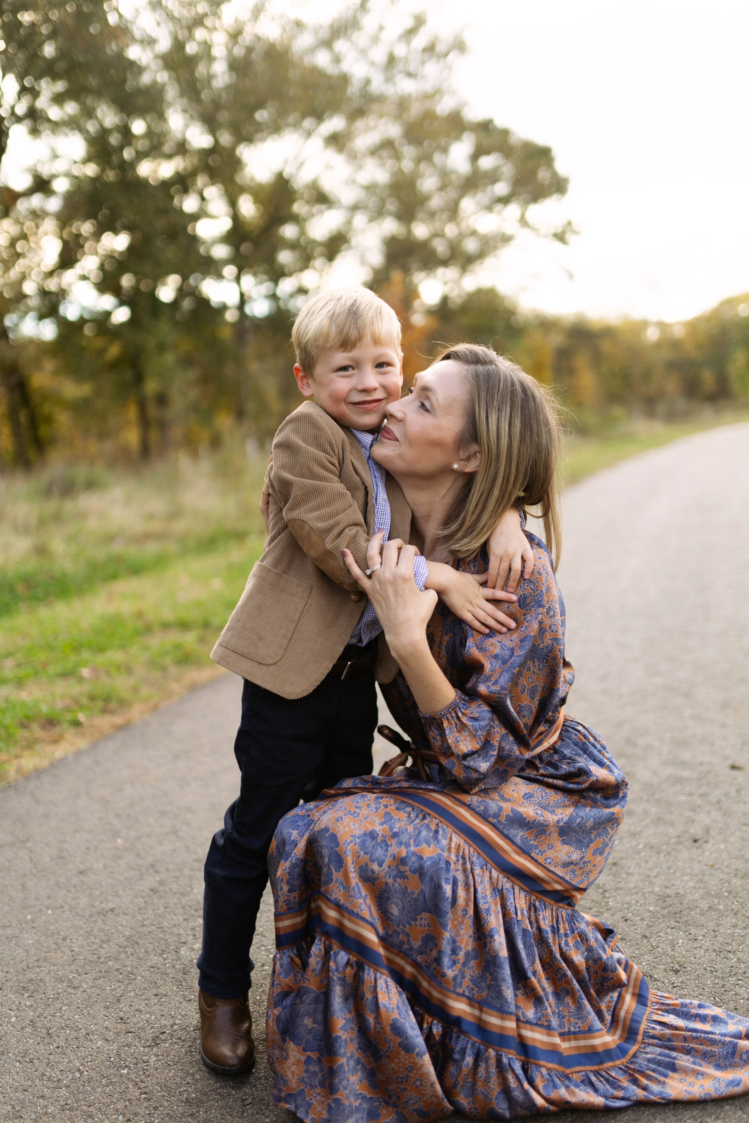 A woman kneeling on the ground and hugging a young boy, both smiling, outdoors in a park during autumn.