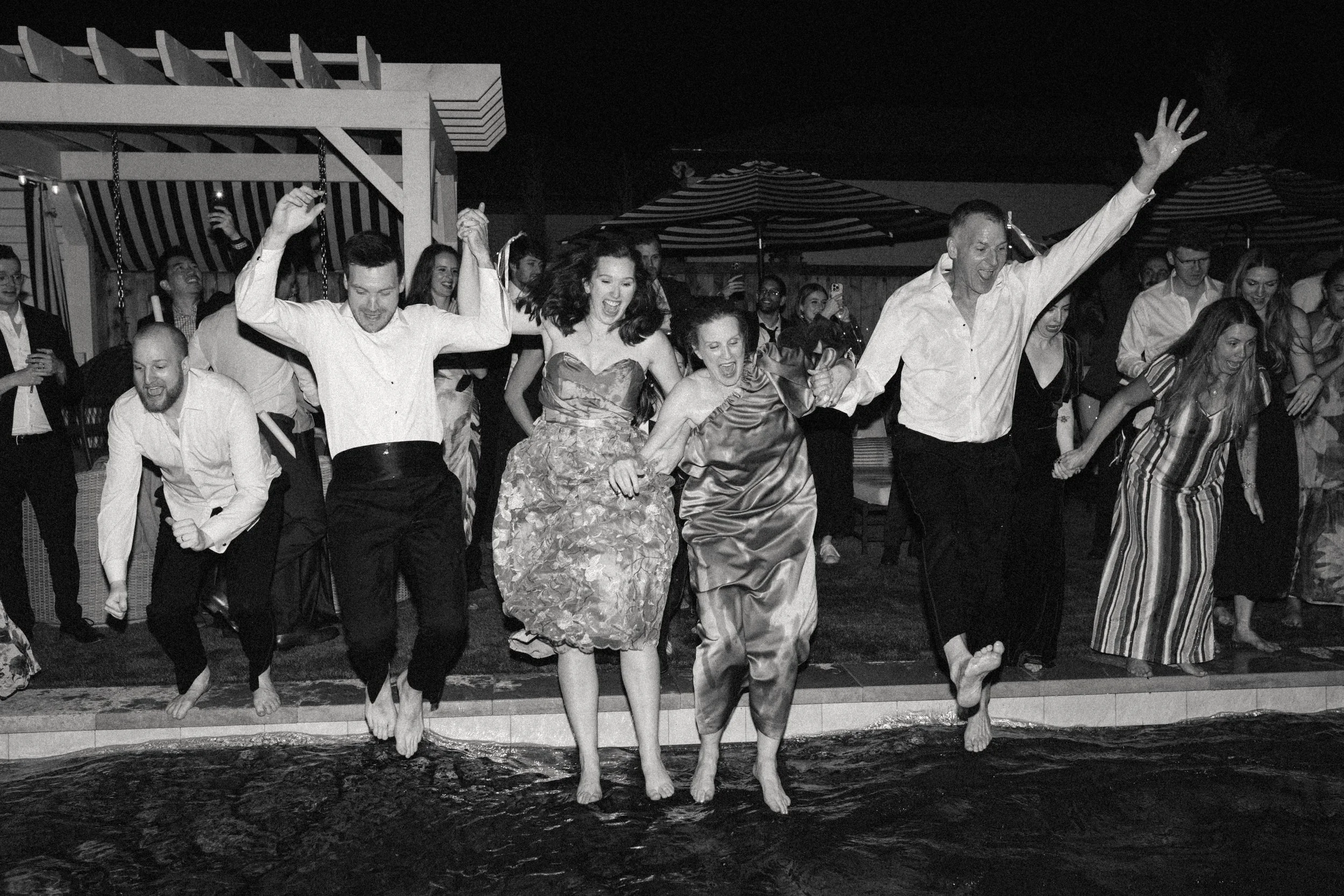 Group of people in formal attire jumping into a pool during a celebration or party at night.