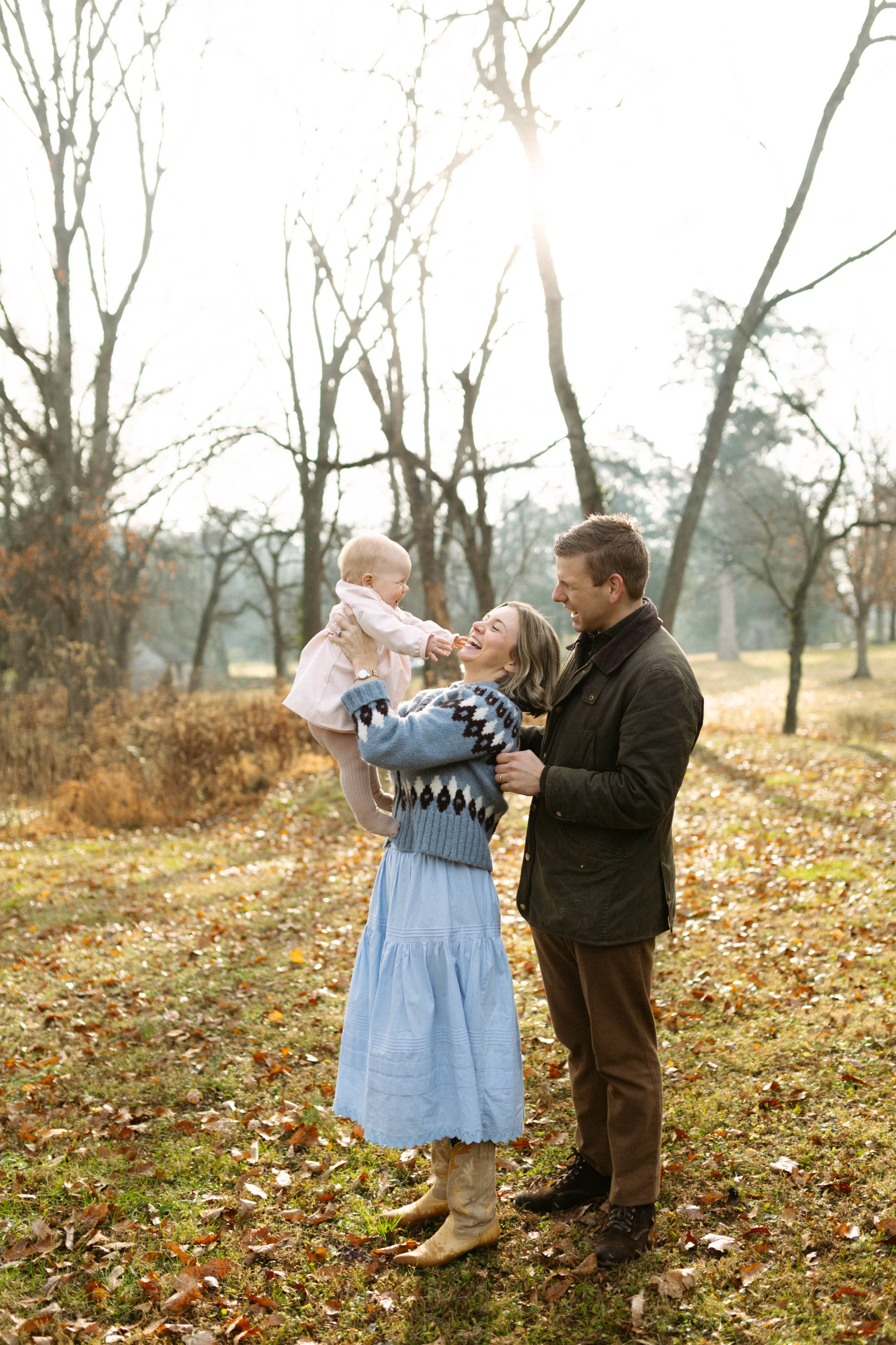 Family of three enjoying outdoor time in a park during fall, with trees and fallen leaves in the background. The woman is holding a smiling toddler girl, while the man stands beside them.