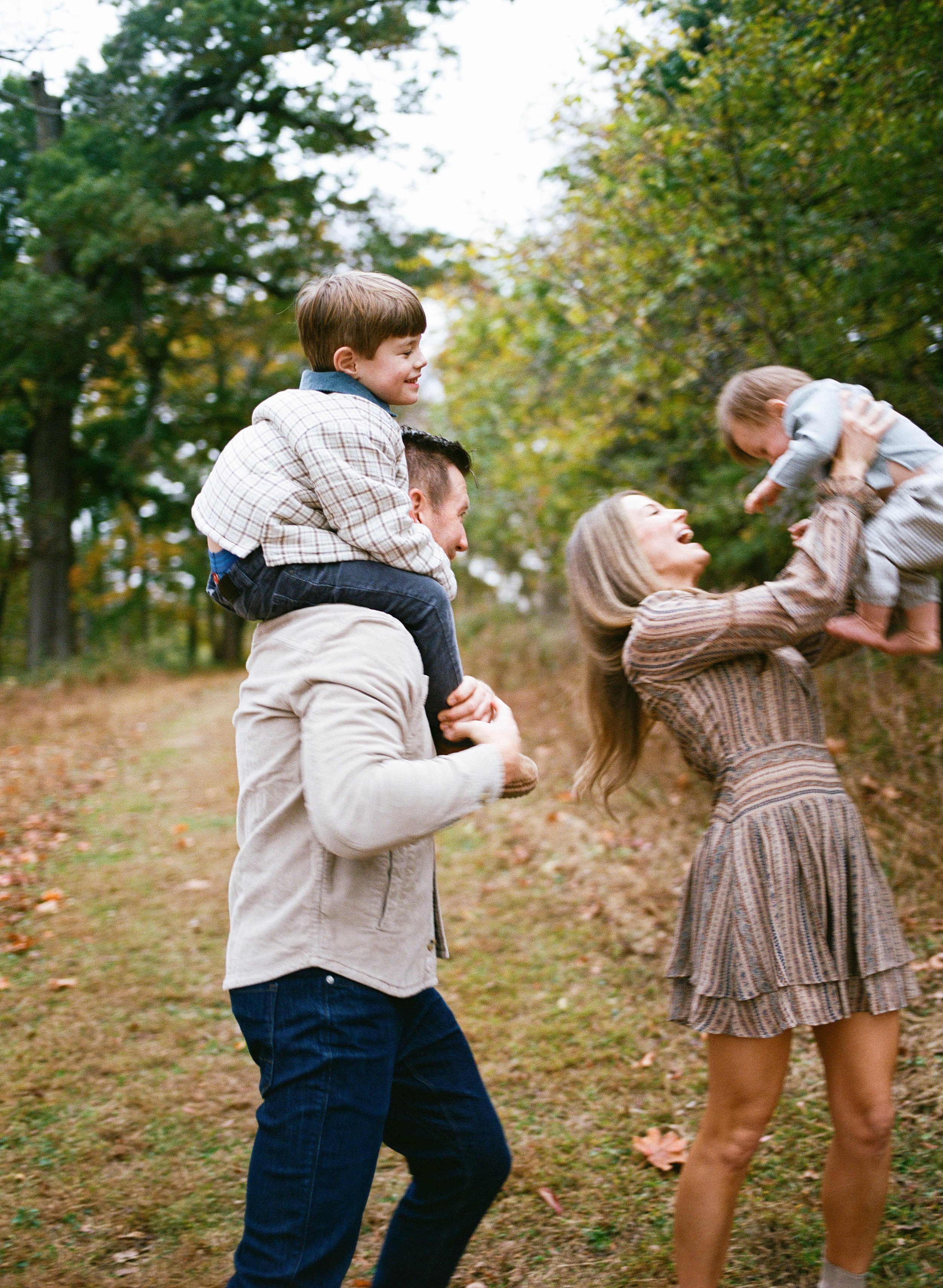 A family enjoying an outdoor moment in a park with trees. The father is carrying a young boy on his shoulders, and the mother is holding a small child in the air, smiling and laughing.