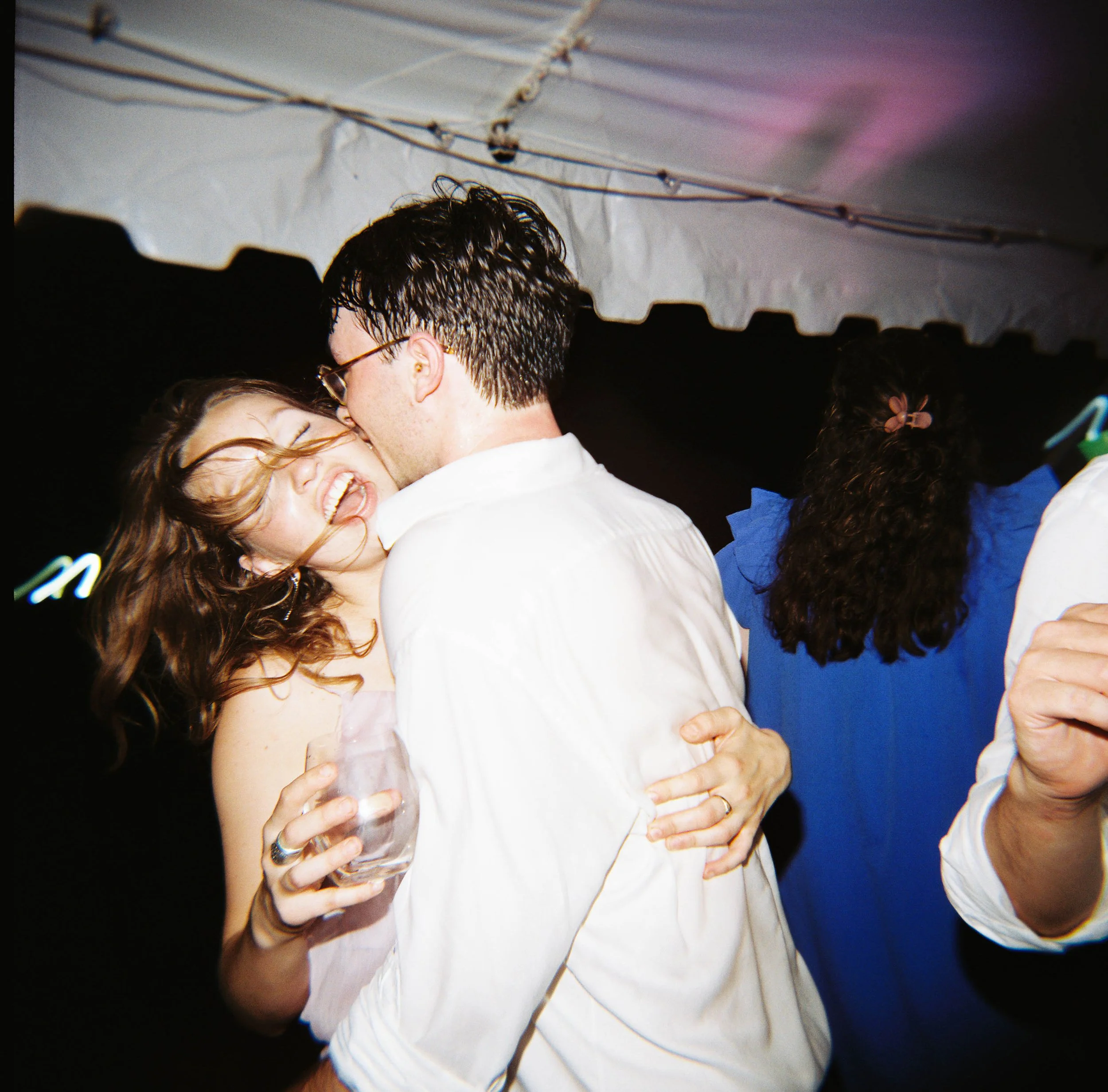 A woman in a green satin strapless dress appears to pour a drink on a man dancing on the floor at a party. The man, wearing a suit and a backward baseball cap, is on the ground with a happy expression. Other people are dancing and socializing in the background.