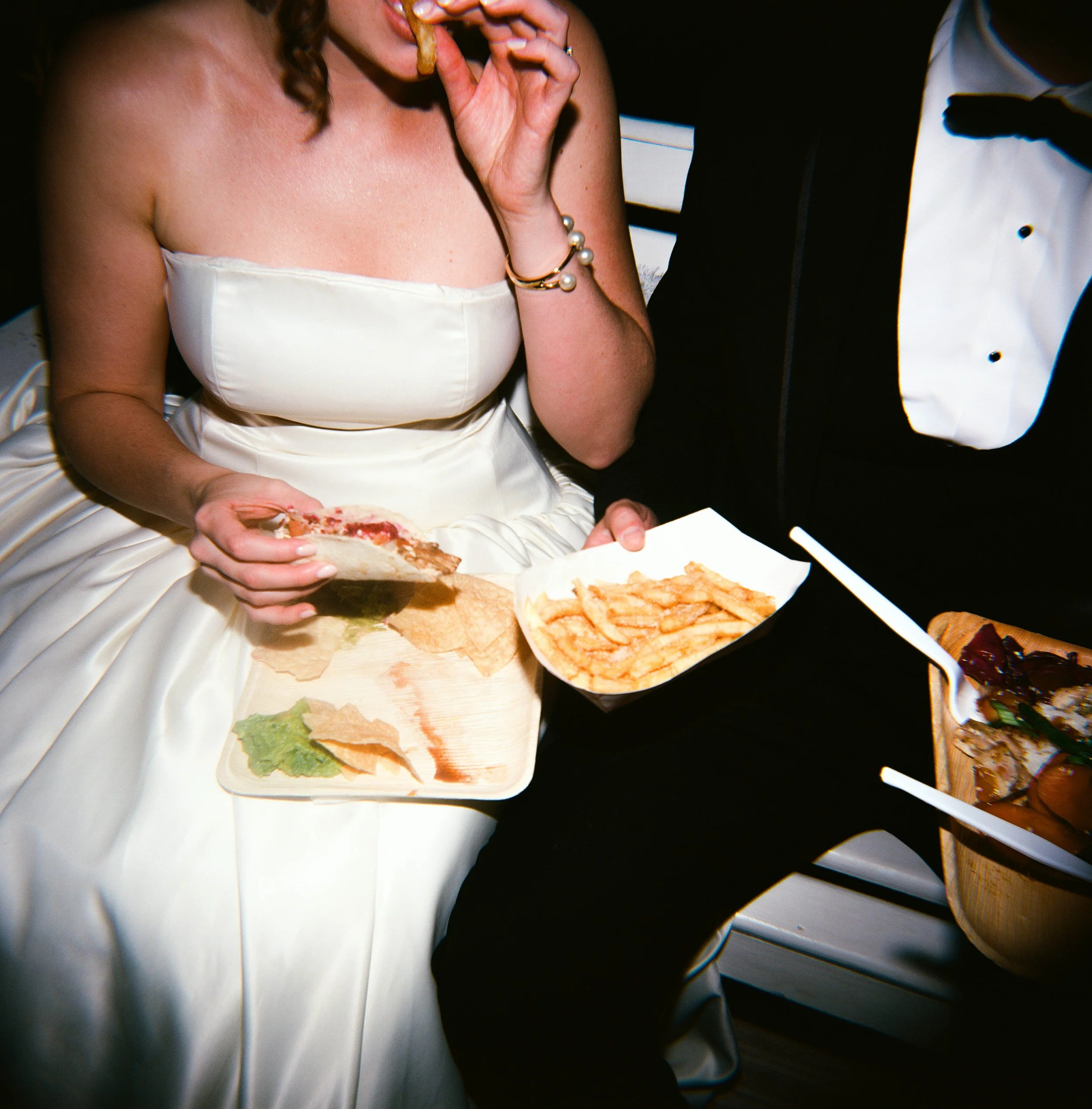 A woman in a strapless white dress eating a slice of pizza, sitting next to a man in a tuxedo, with various food dishes and fries on a table.