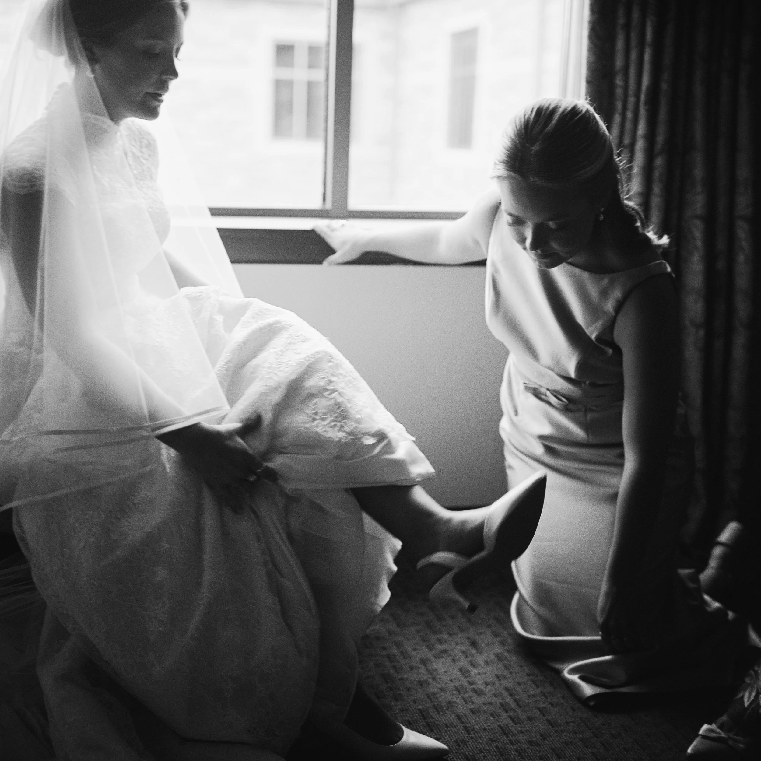 Two women, one in a wedding dress and the other in a sleeveless dress, are sitting on the floor near a window with natural light coming in, engaged in an emotional moment.