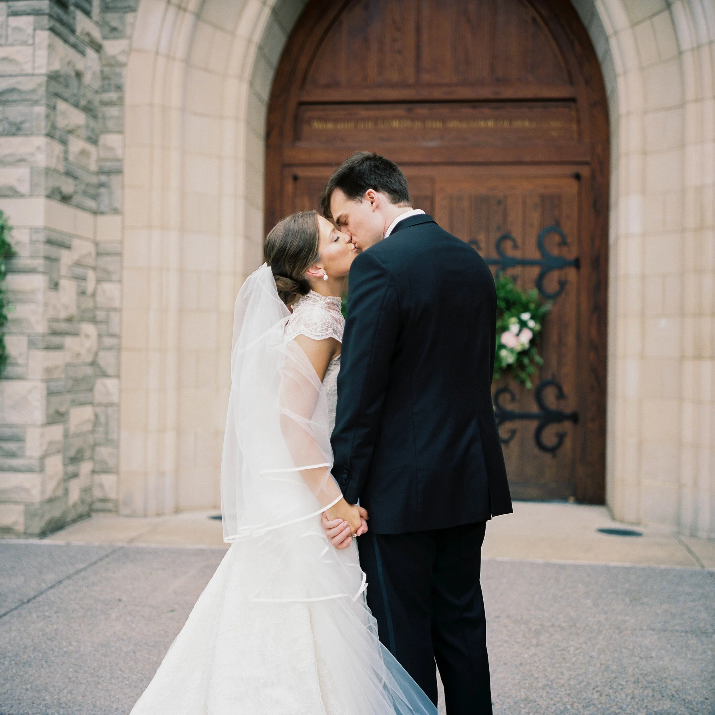A bride and groom sharing a kiss in front of a church door, holding hands, with the bride wearing a white lace wedding gown and veil, and the groom in a dark suit, during their wedding ceremony.