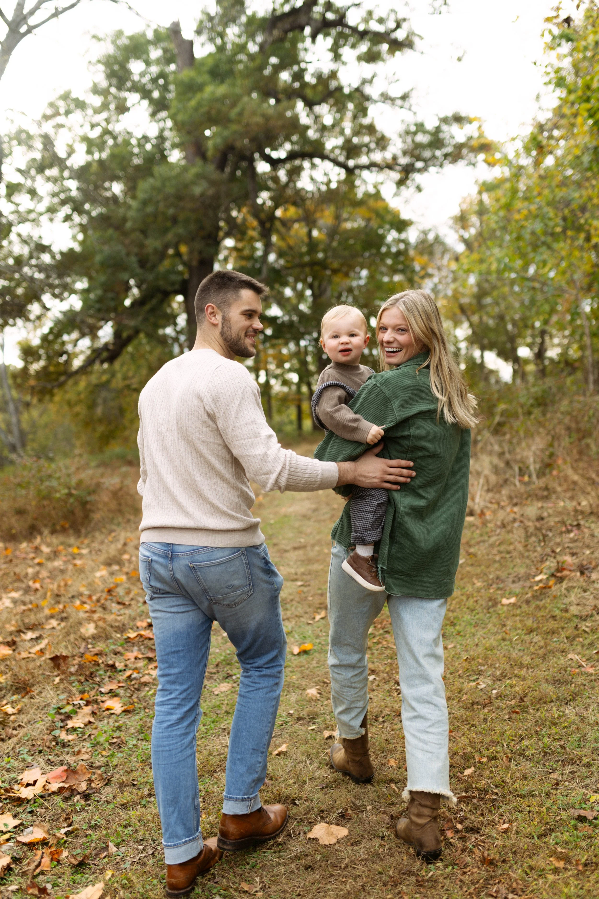 A happy family of three, a man and woman with a small child, walking outdoors in a wooded area with autumn leaves on the ground.