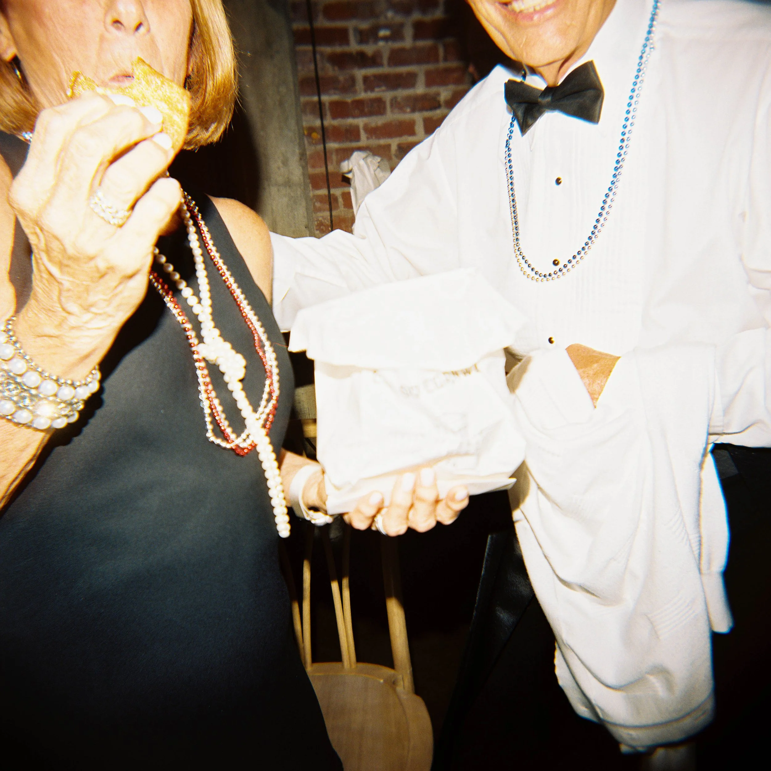 Two people at a social gathering, smiling and holding each other's hands. One person is wearing a tuxedo with a bow tie and a beaded necklace, and the other is wearing a black dress with pearl necklaces. They appear to be enjoying a special moment to