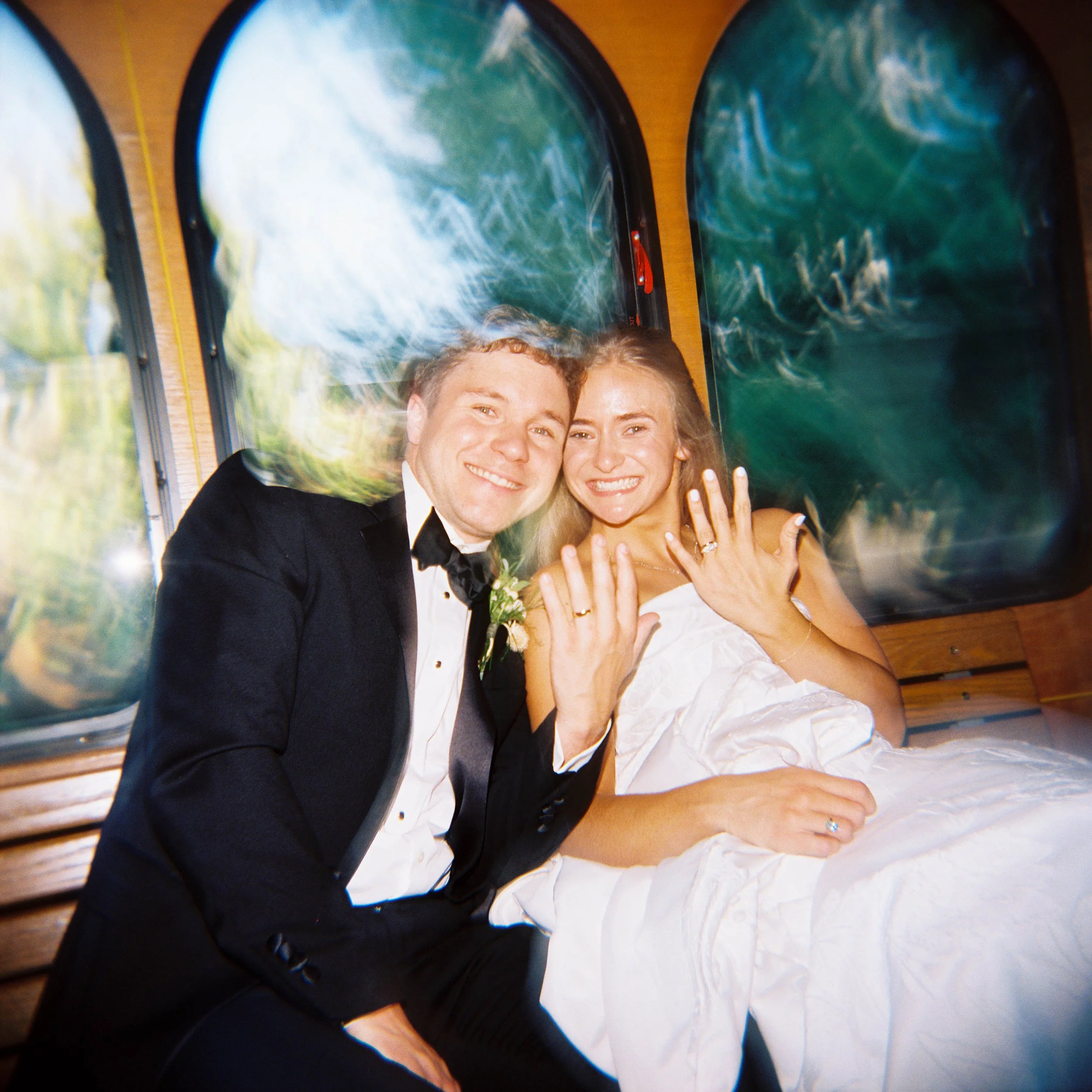 A smiling couple in wedding attire, the man in a tuxedo and the woman in a white wedding dress, posing happily in front of a backdrop of water with a wooden table surface.