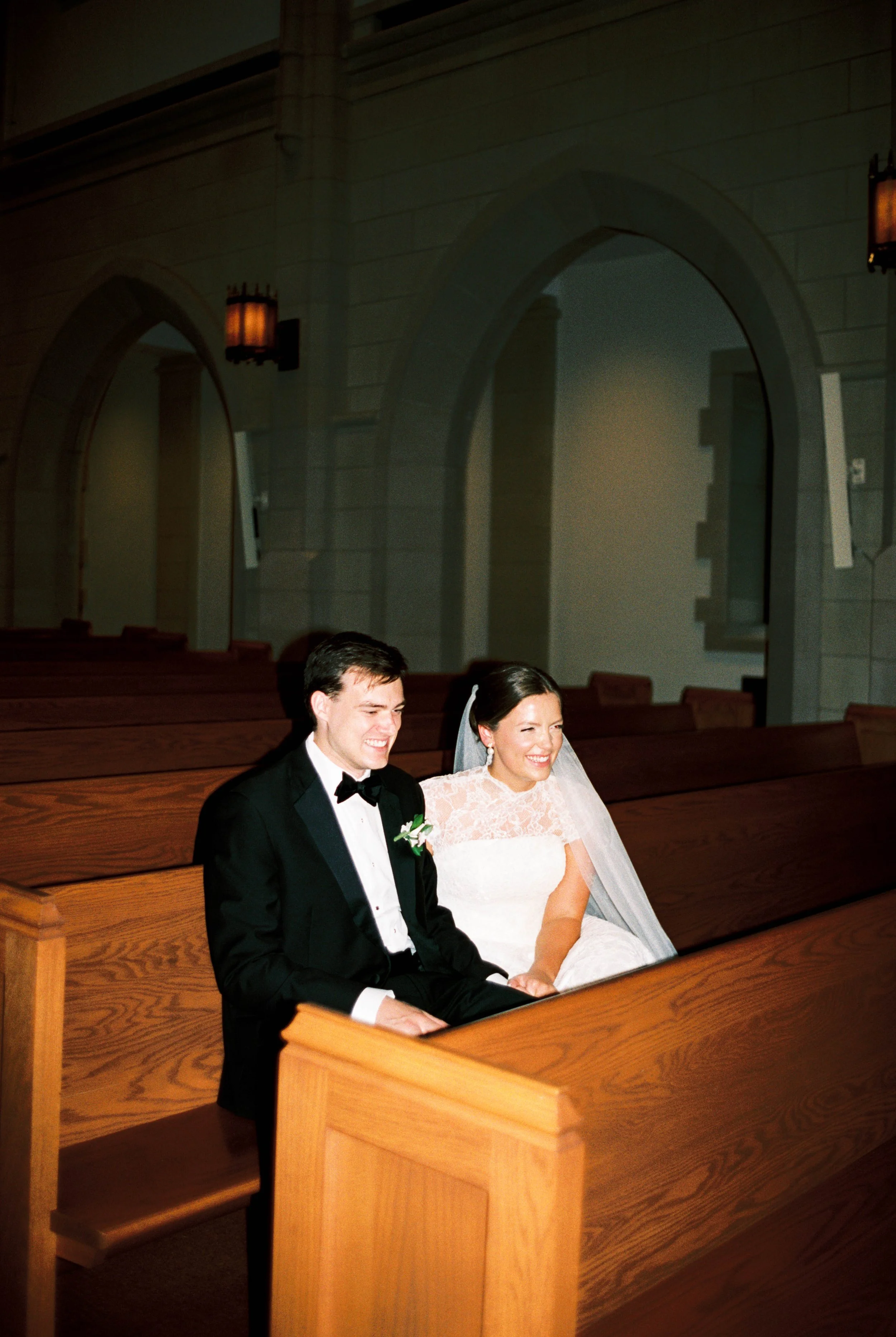 A bride and groom sitting on a church pew during their wedding ceremony, smiling and looking happy.