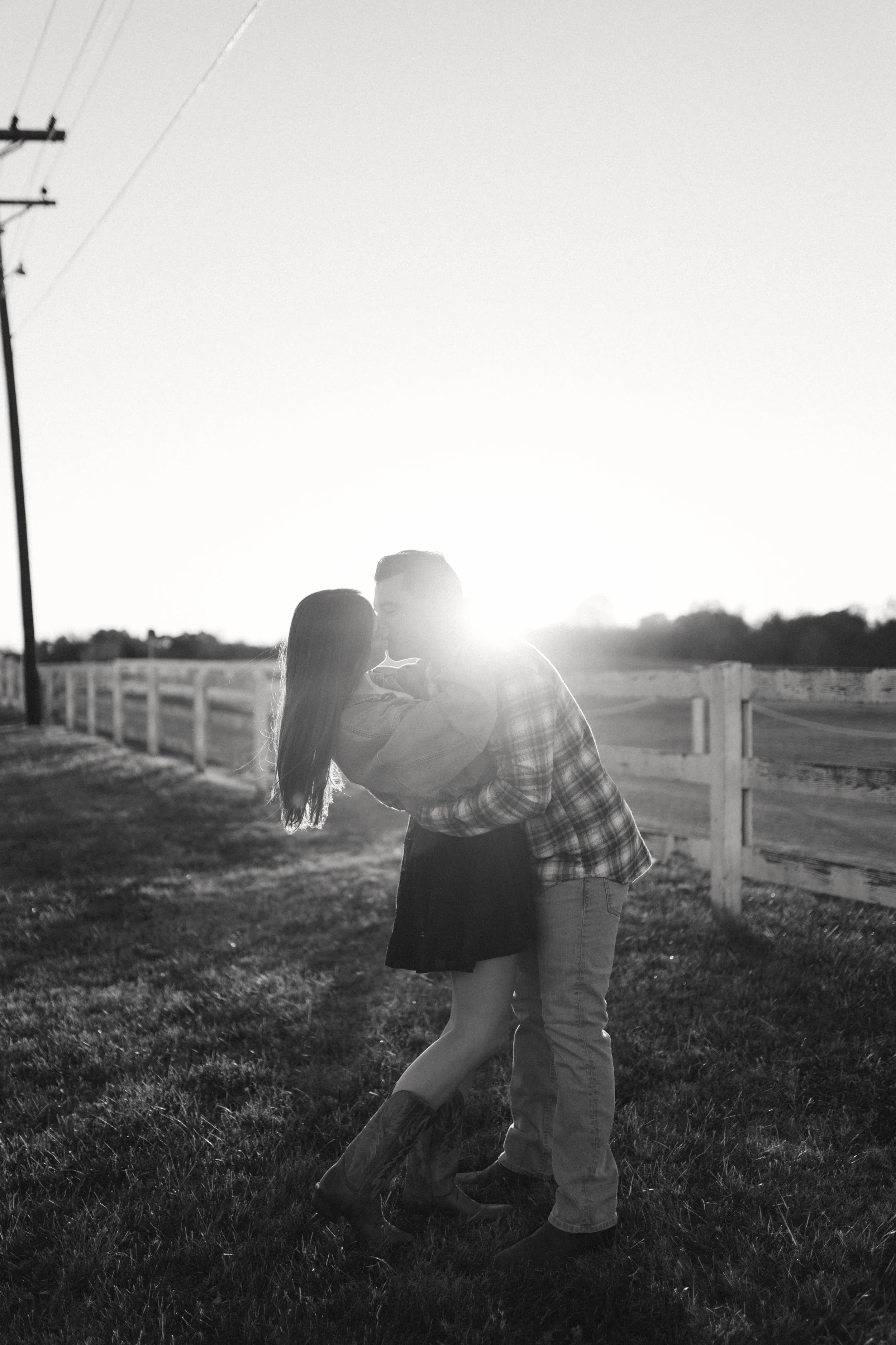 A black and white photo of a couple kissing outdoors with the sun setting behind them, on a grassy area next to a white wooden fence.