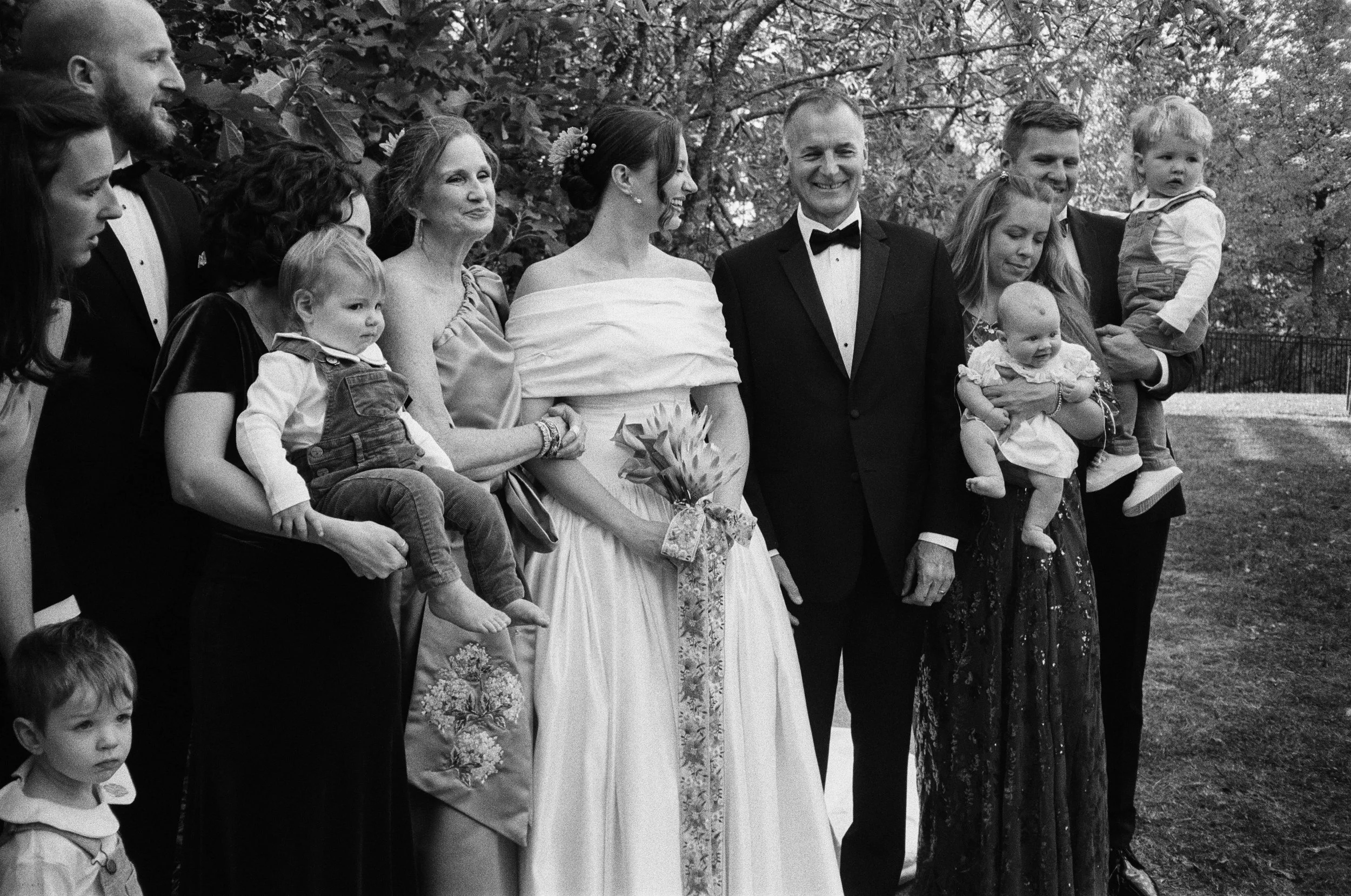 Group of people gathered outdoors, including a bride and groom, holding children and standing on grass with trees in the background, at a wedding.