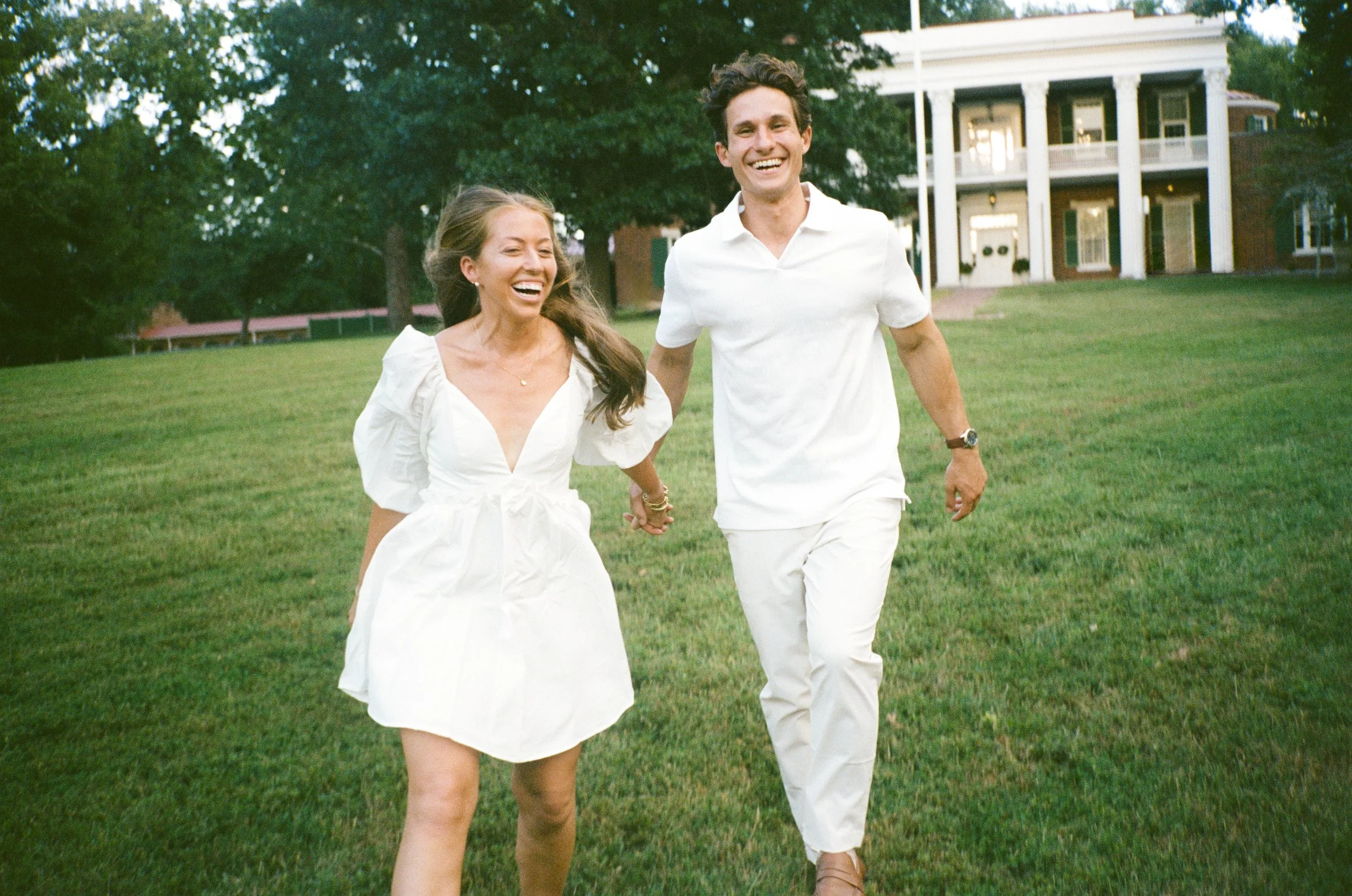 A smiling young woman and man walking hand in hand across a grassy field with a large white house in the background.