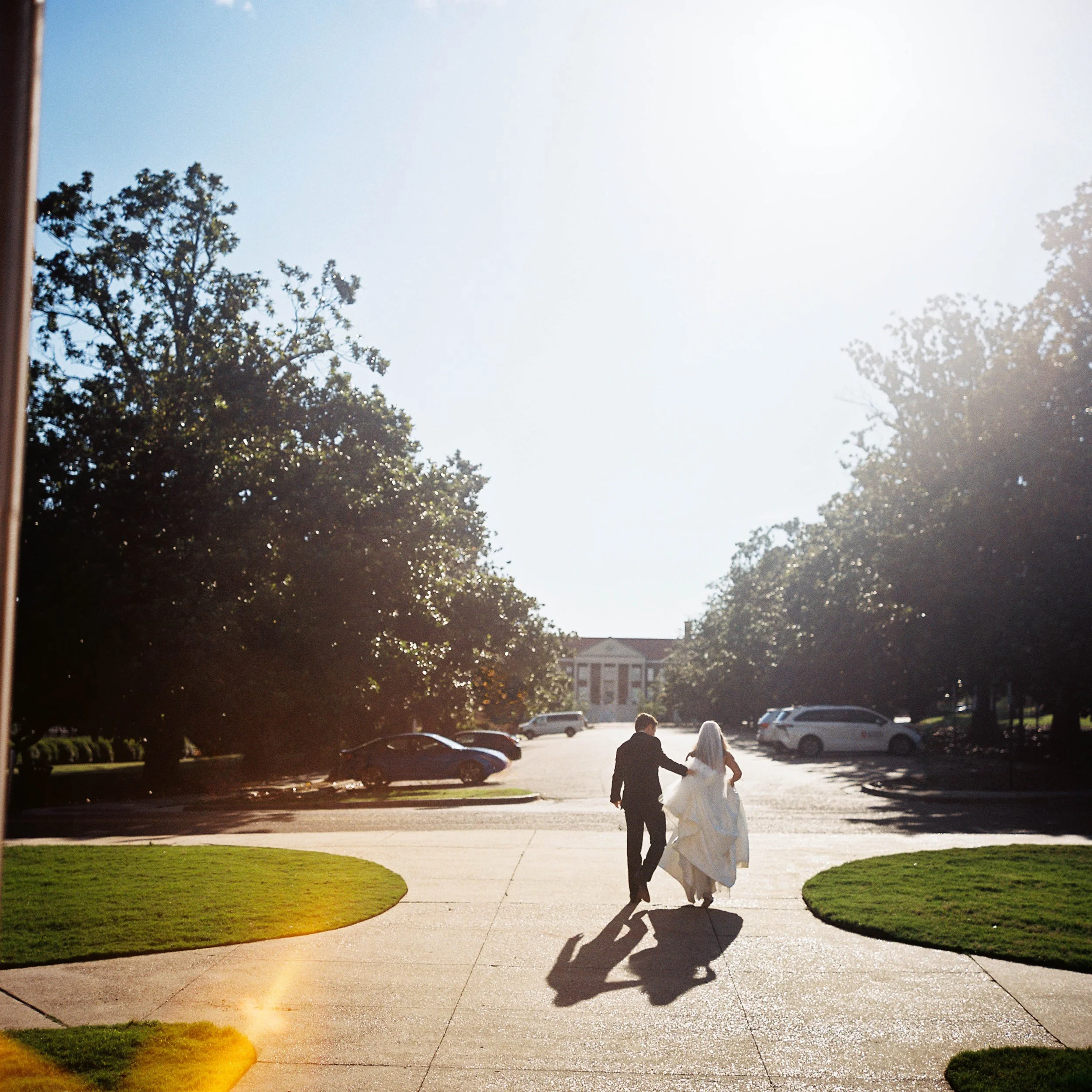A bride and groom walking hand in hand on a sidewalk in front of a parking lot with cars and a building in the background. The sun is shining brightly, creating shadows and a lens flare effect.