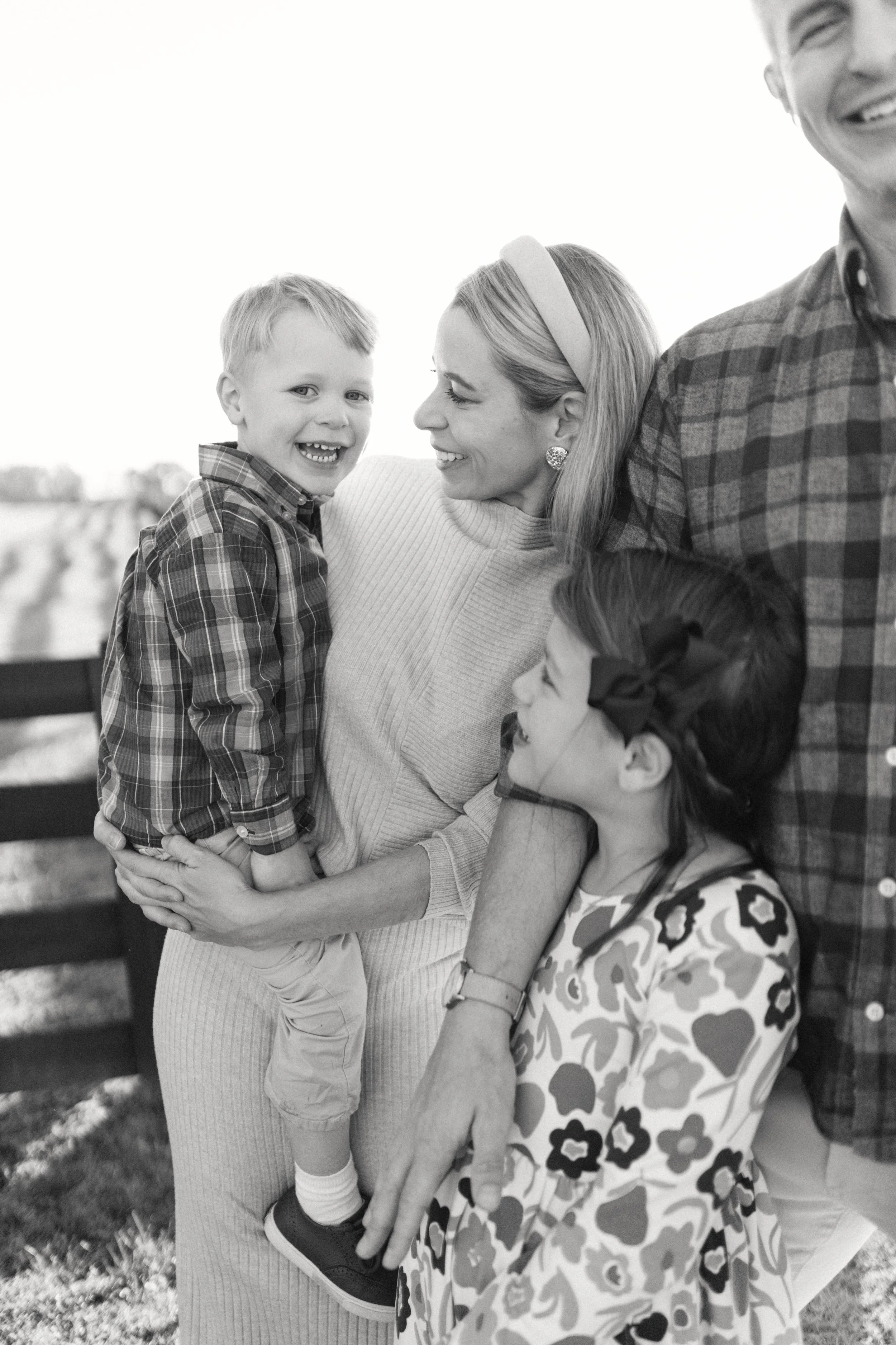 A smiling woman holding a young boy in her arms, with a girl beside her and a man in a plaid shirt, outdoors in a rural setting.