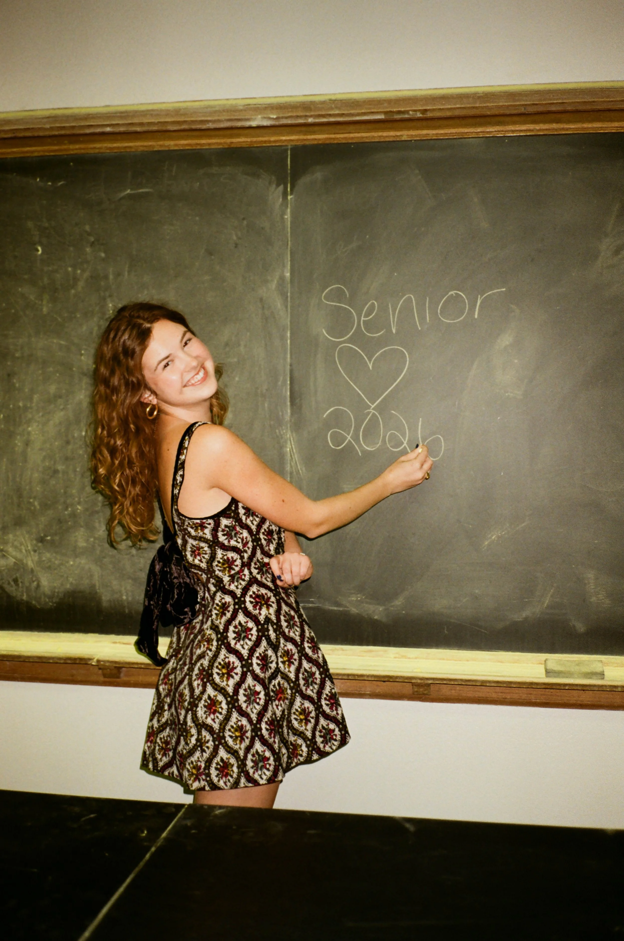 A young woman with curly brown hair and a patterned dress is writing on a chalkboard, which says 'Senior ♥ 2020'.