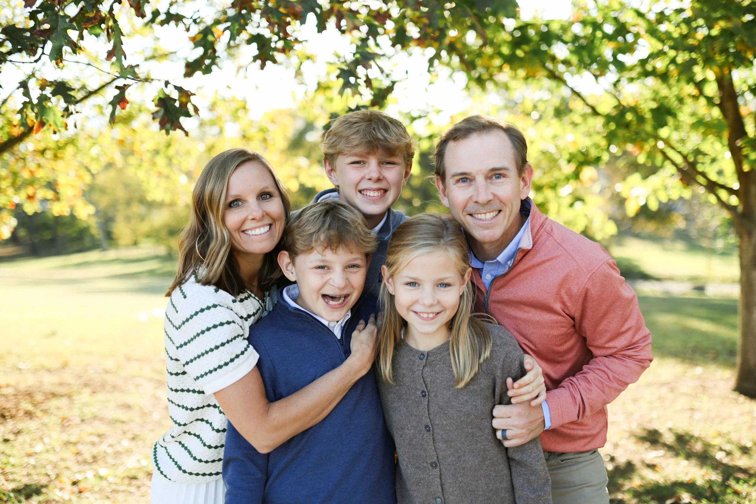 A happy family of six outdoors in a park during fall, with trees and sunlight in the background. The family includes parents and four children, all smiling and closely posing together.