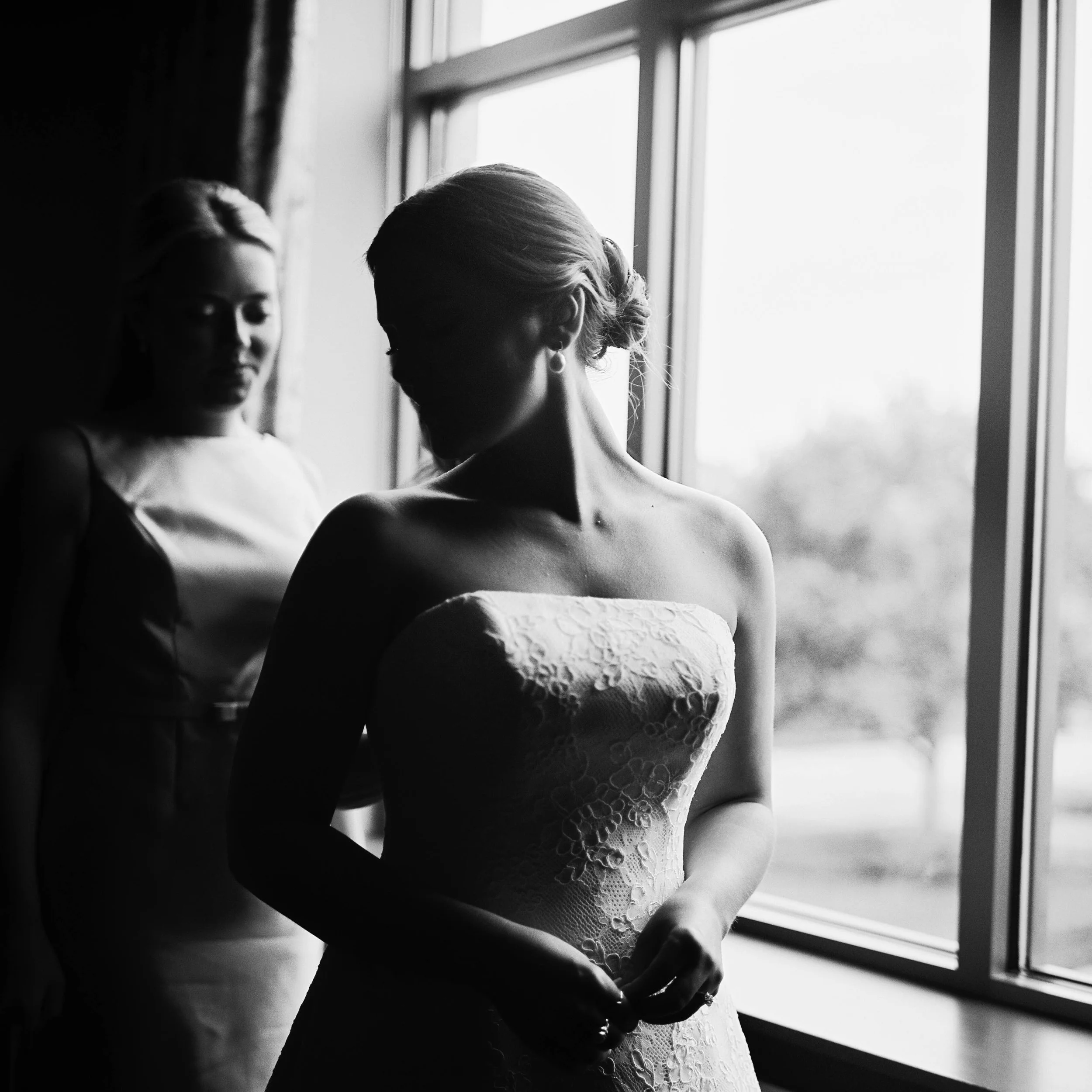 Black and white photograph of a woman in a lace dress looking downward, standing beside a window with light streaming in, creating a silhouette effect.