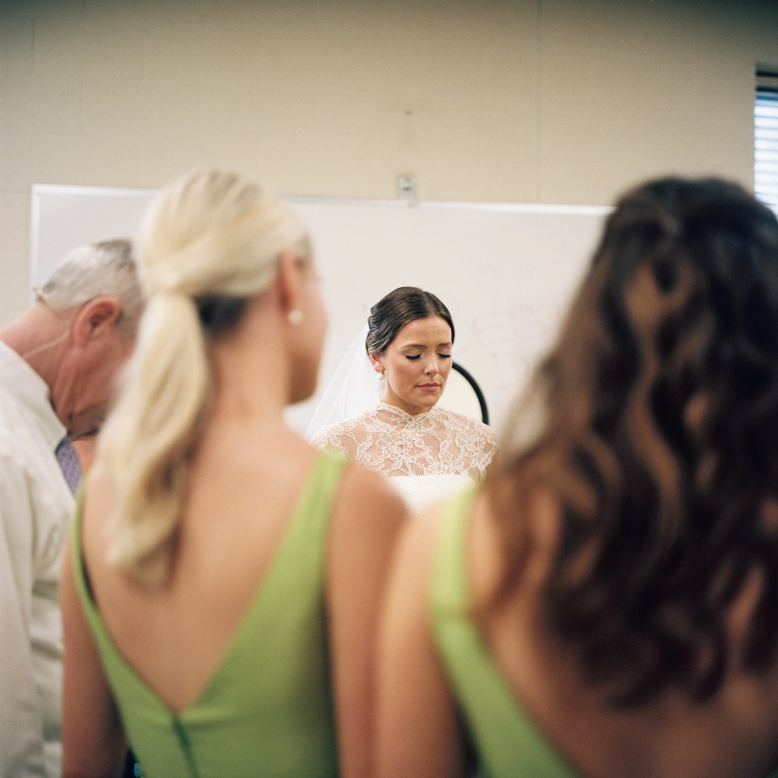 Close-up of a woman with dark hair wearing lace top, surrounded by people with blonde hair and light-colored clothing in a room.