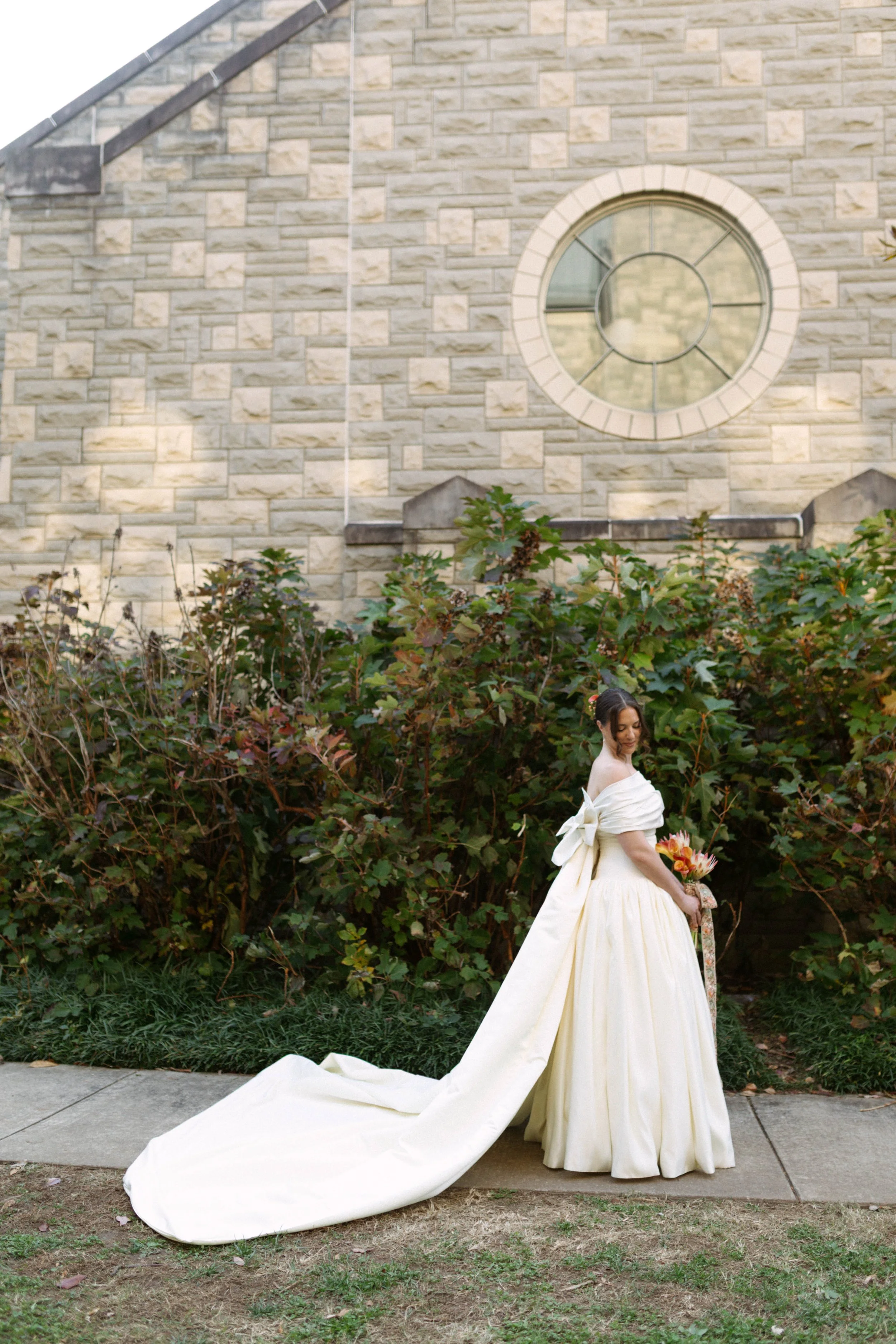 Bride in a white wedding gown with a long train standing outdoors, holding a bouquet of flowers, with a stone building and large circular window in the background.