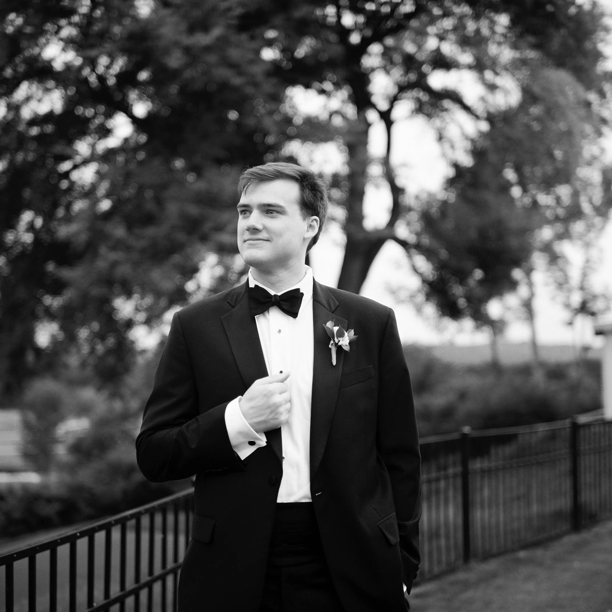 A young man wearing a tuxedo with a bow tie, standing outdoors near a black metal fence and trees in the background, in black and white.