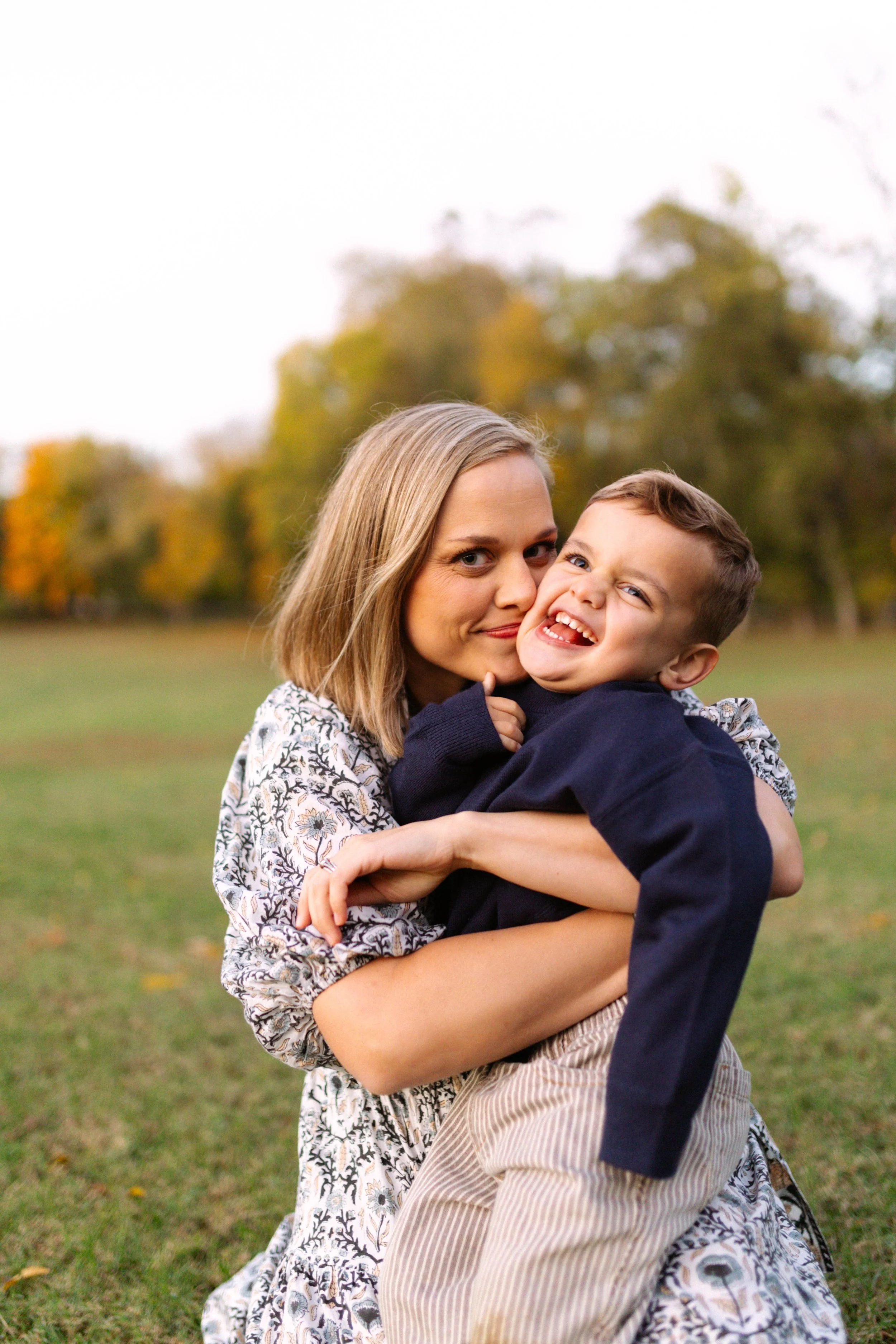A woman and young boy hugging outdoors on a grassy field with autumn trees in the background.