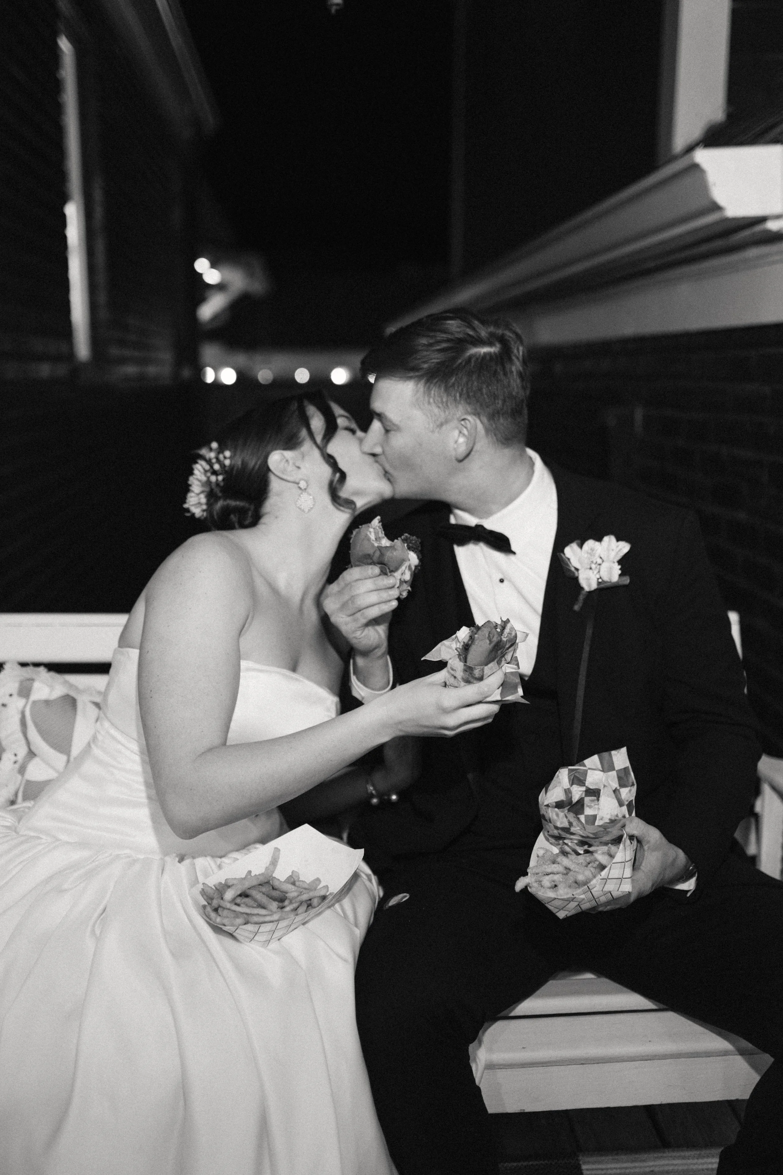 Black and white photo of a bride and groom sharing a kiss while holding food, sitting on a bench at night, with fries and burgers in paper trays.