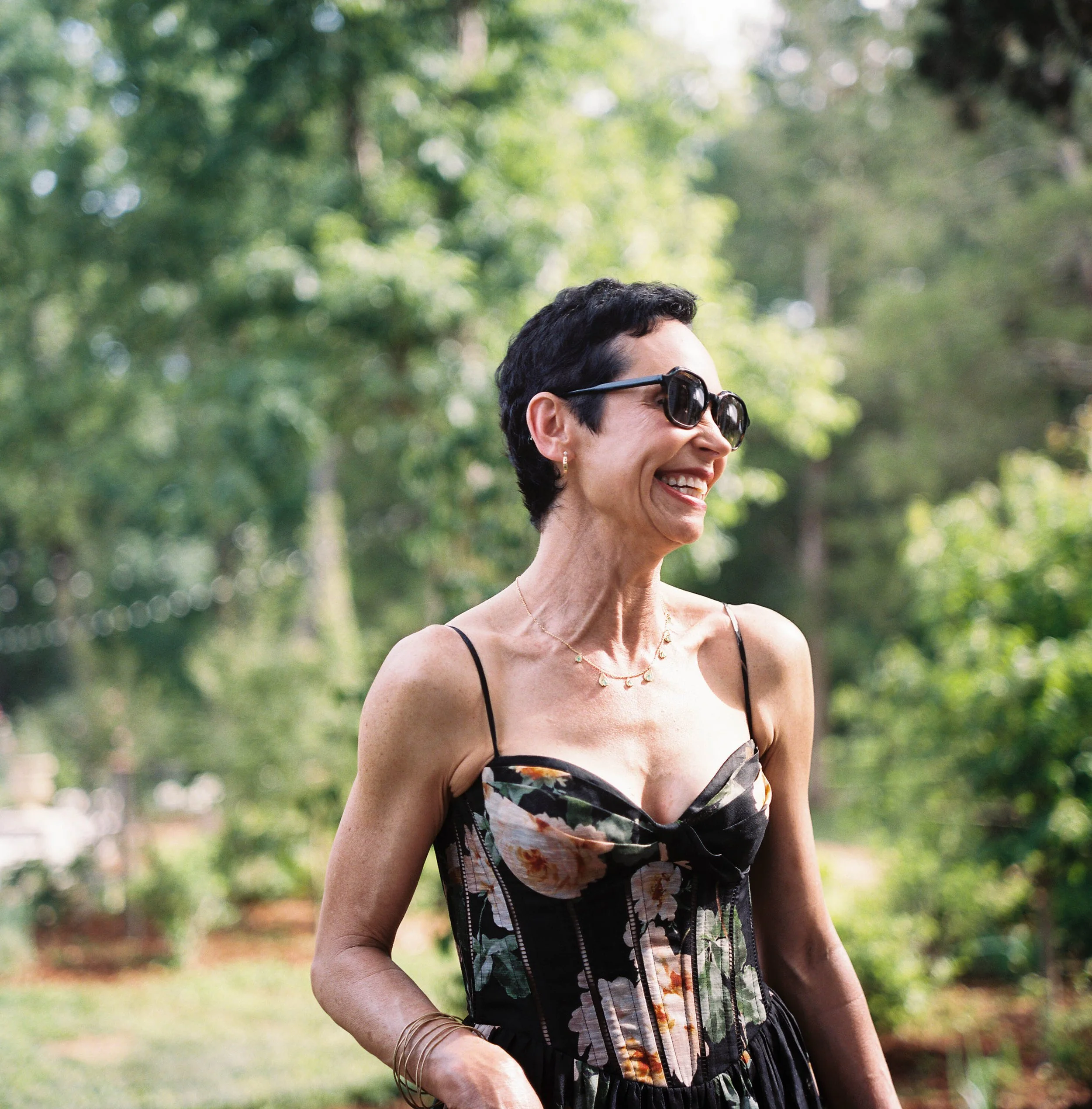Smiling woman wearing sunglasses and a floral and striped dress outdoors in a green park.