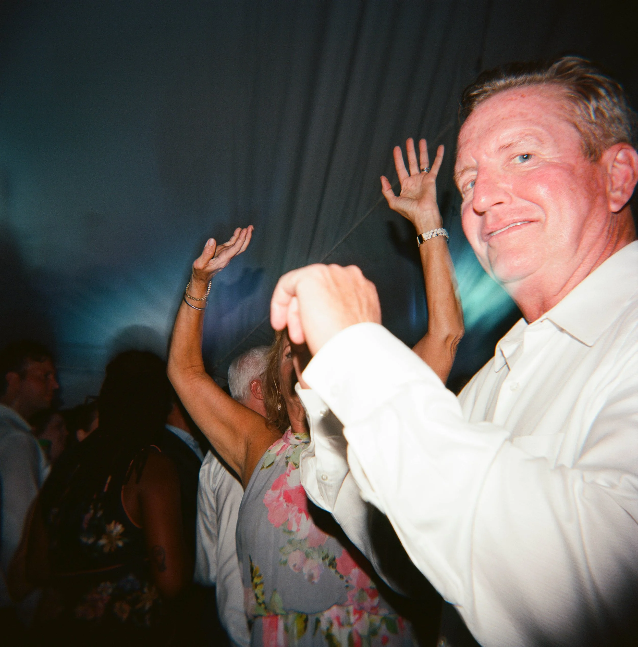 Close-up of a man in a white shirt with pink floral pattern, smiling, with several hands raised around him, some touching his head and shoulder, in a crowded setting.