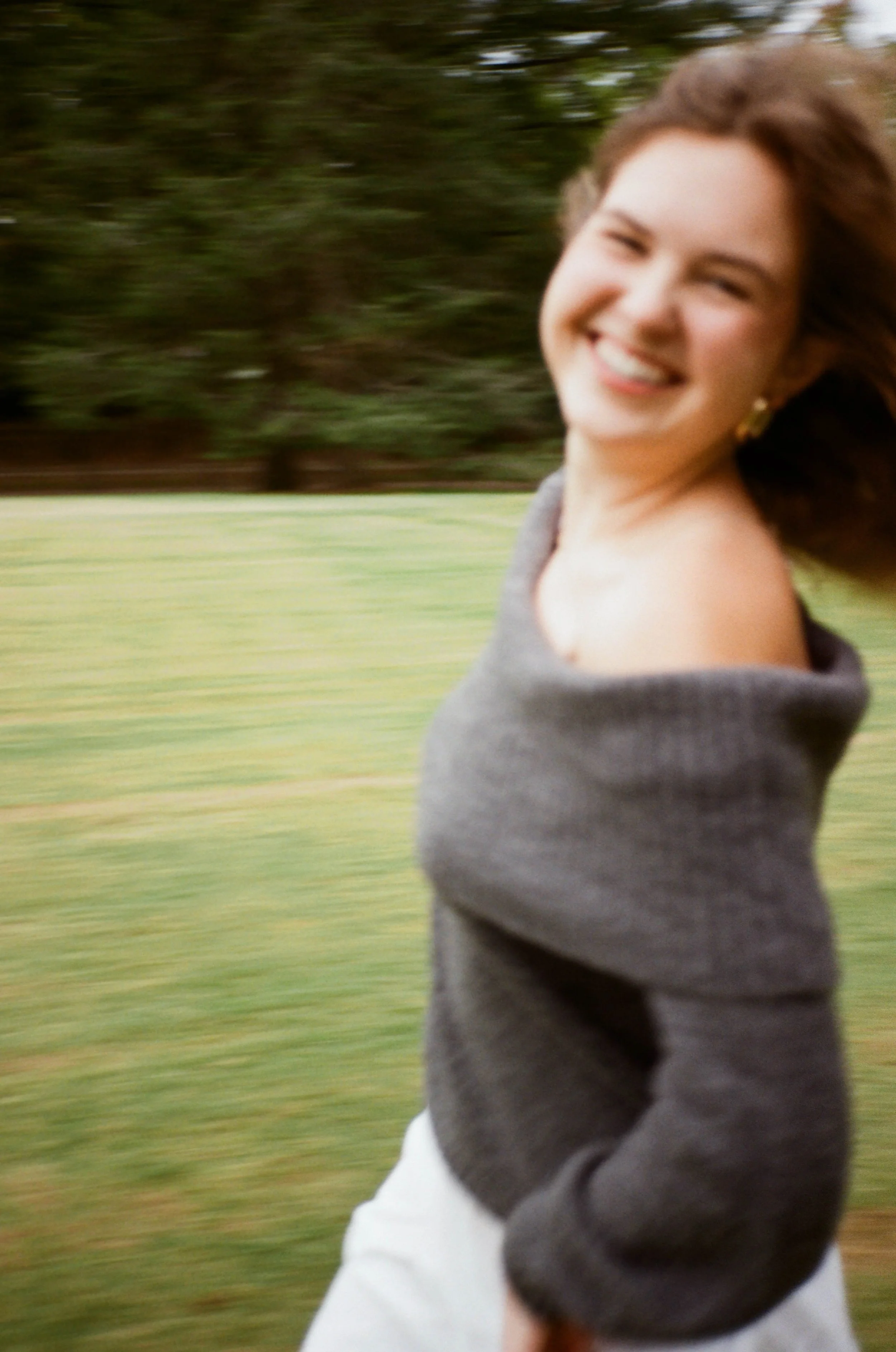 A smiling woman with brown hair and gold earrings running outdoors on a grassy area, wearing a gray off-the-shoulder sweater.
