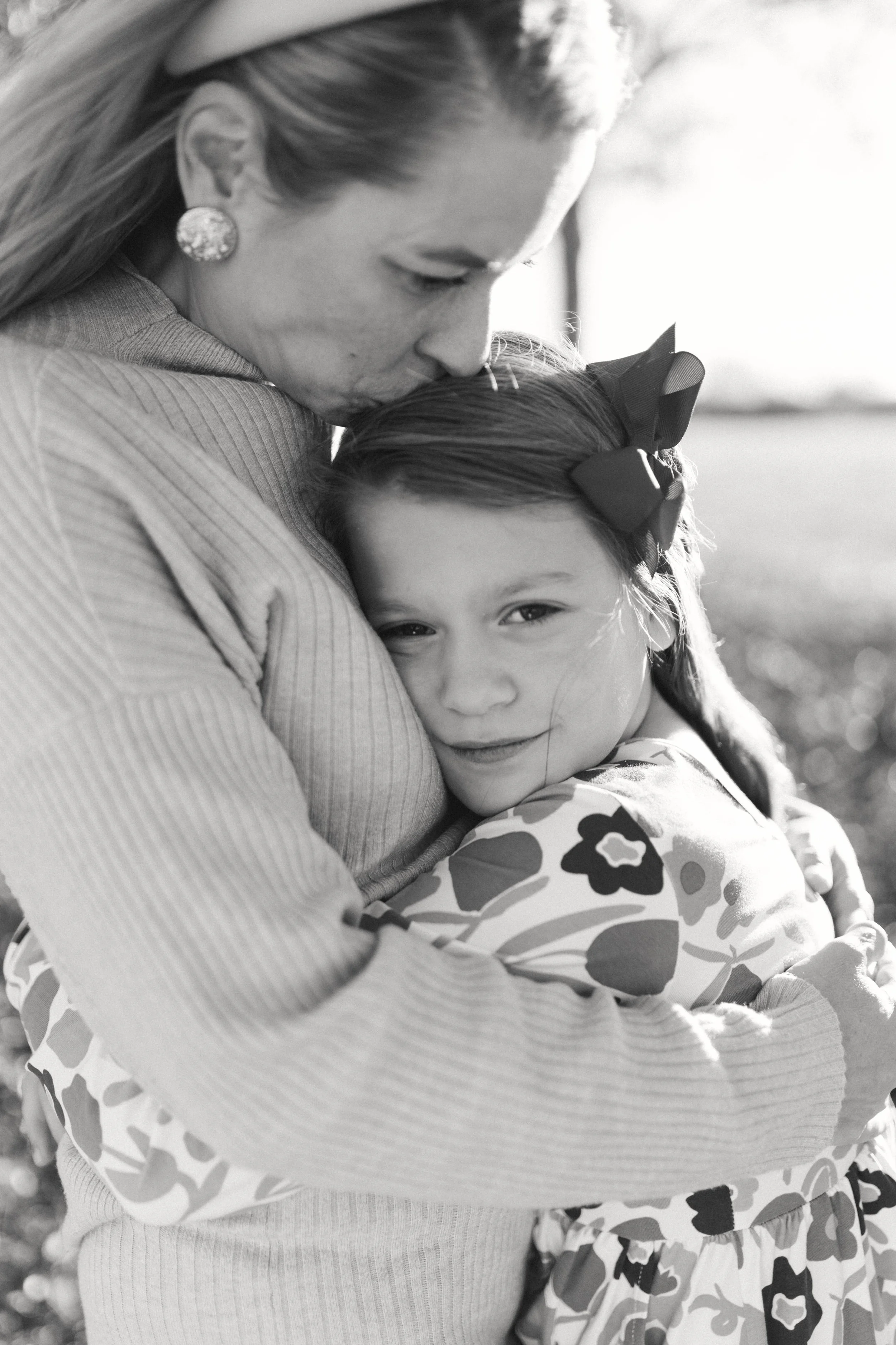 A woman holds a young girl in a hug outdoors. The girl has a bow in her hair and a patterned dress. The woman is wearing earrings and a ribbed sweater. The scene is backlit, creating a soft glow and a bokeh background.