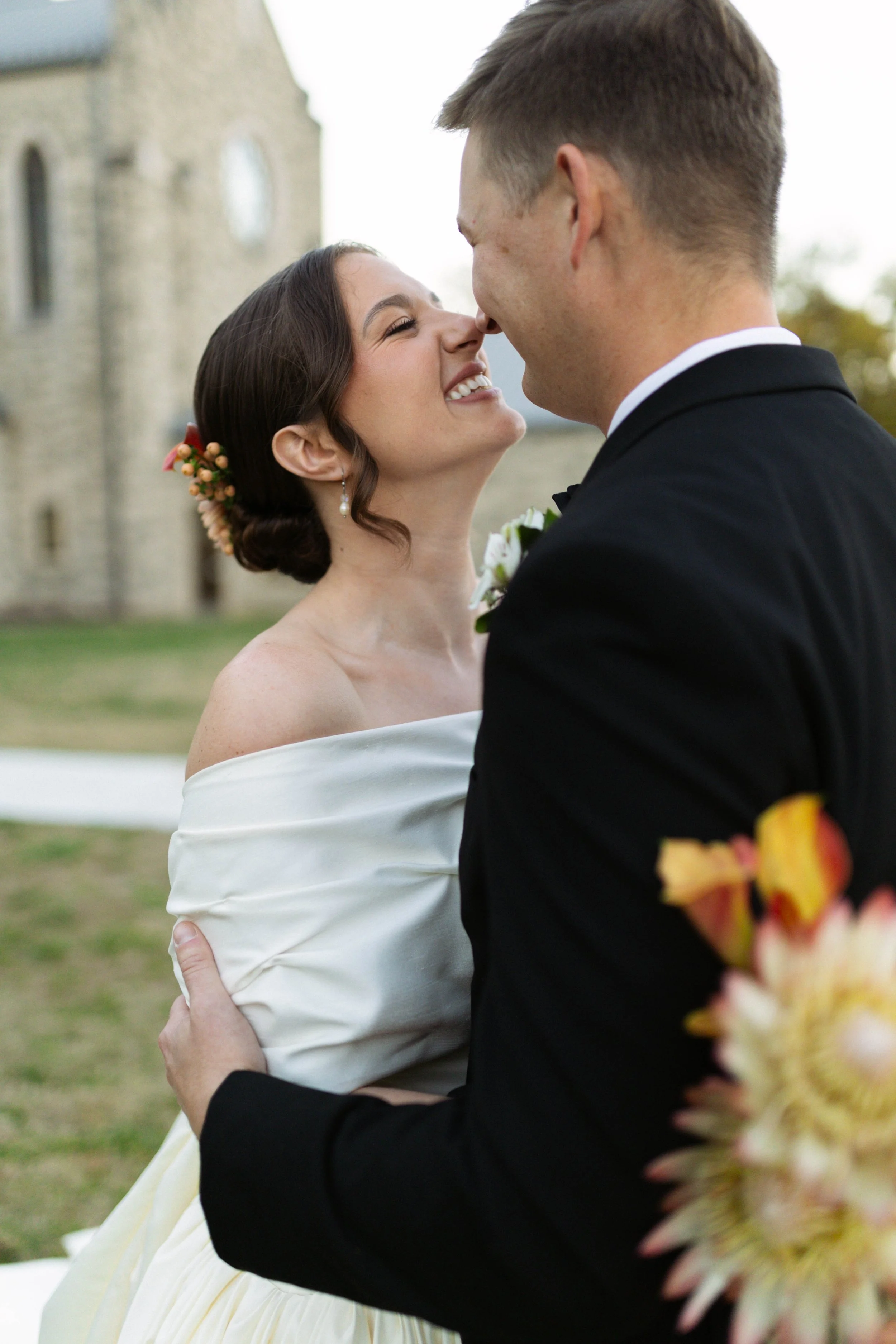 A bride and groom share a close moment outdoors in front of a stone building, smiling and about to kiss.