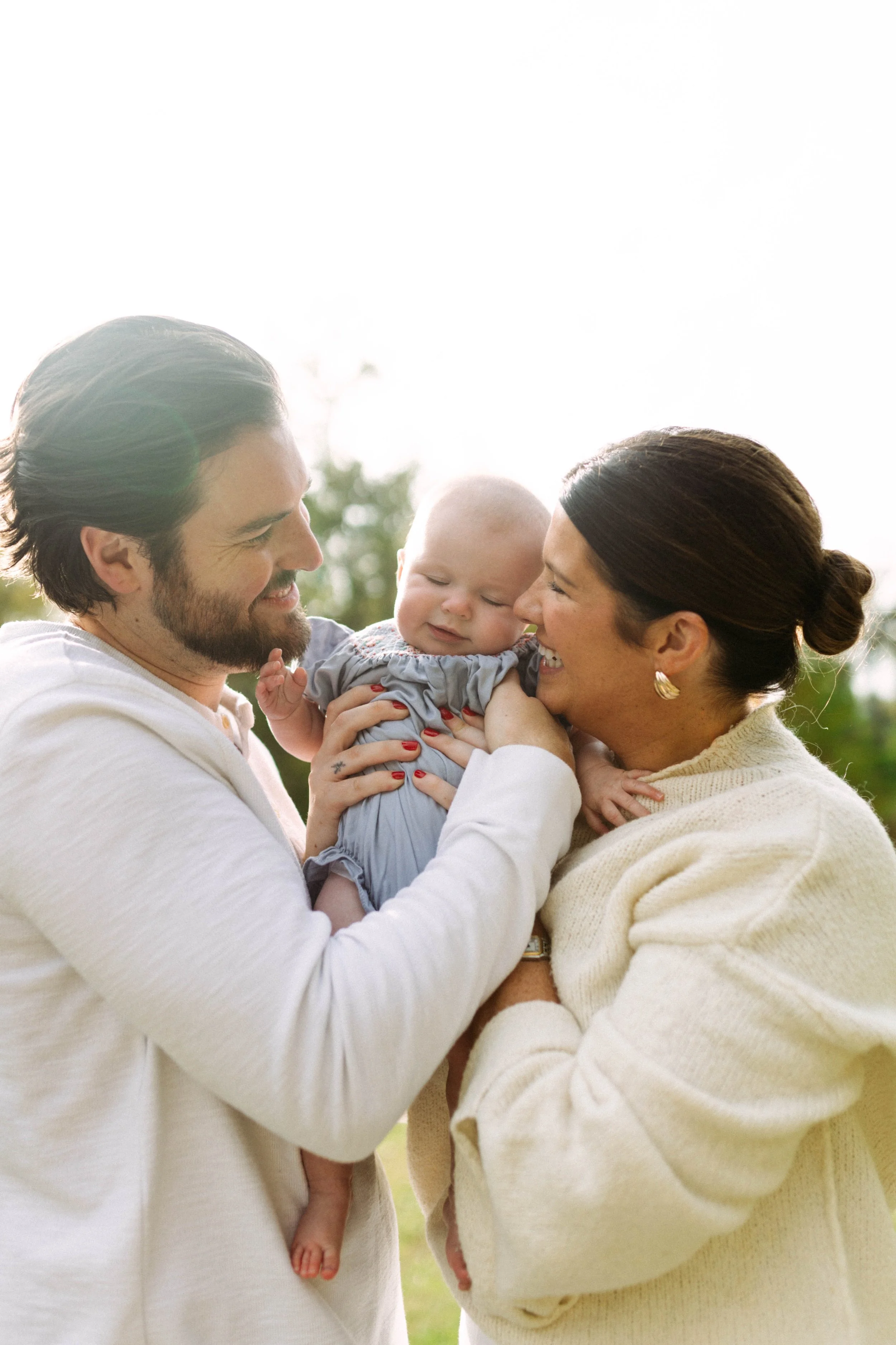 A smiling man and woman holding a baby outdoors, with trees and sky in the background.