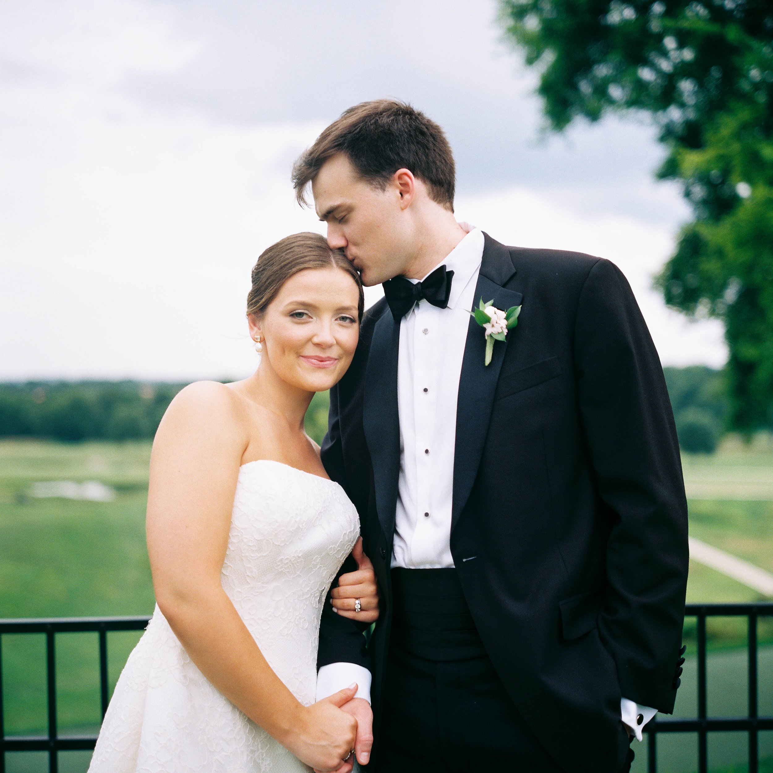 A bride and groom sharing a tender moment outdoors, with the groom kissing the bride on the forehead, while she smiles softly, dressed in wedding attire, with trees and a cloudy sky in the background.