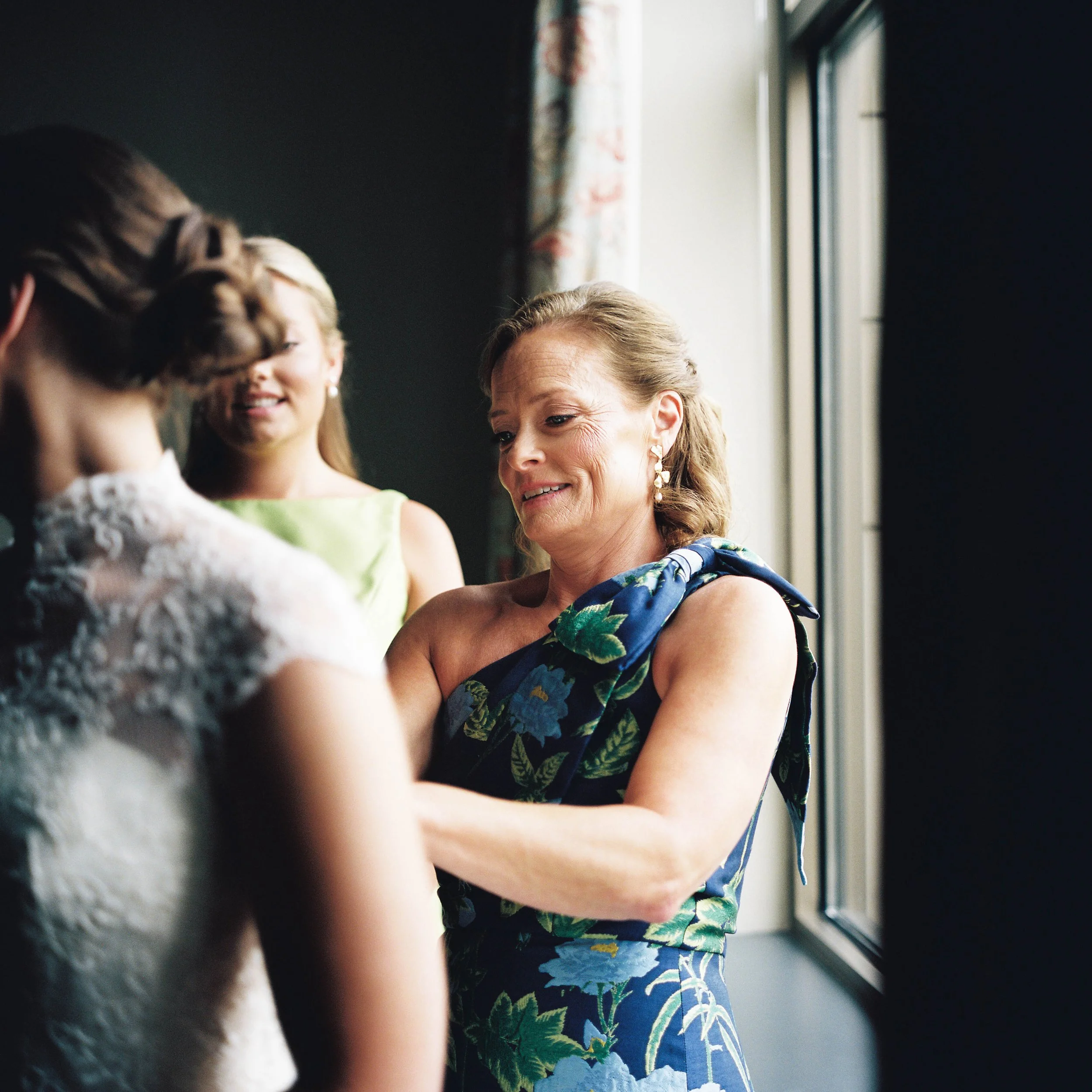 A woman with her hair styled in waves is smiling as she interacts with two children, one wearing braids and the other with light-colored hair, near a window with floral curtains.