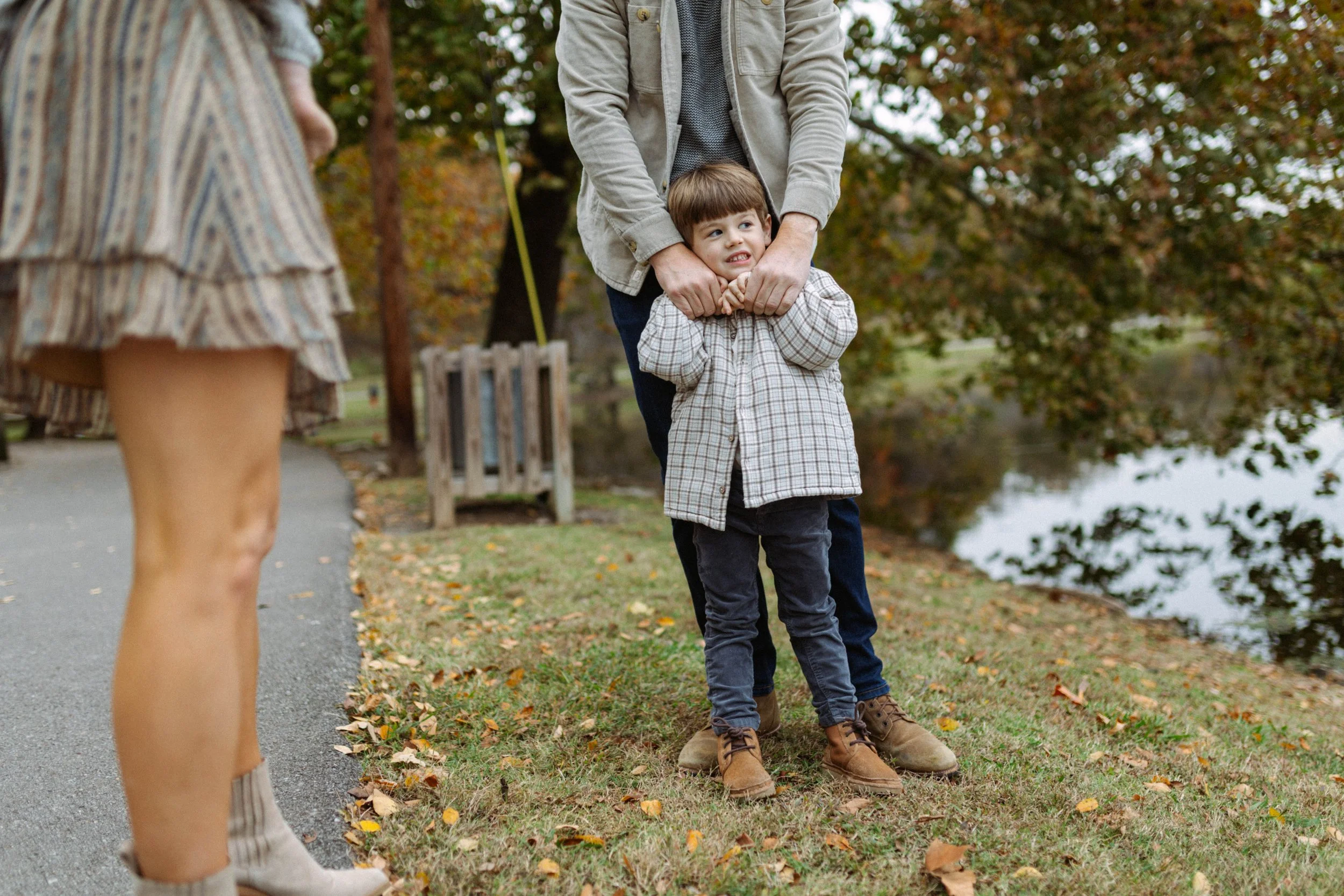 A young boy is being playfully restrained by an adult male, possibly his father, near a lake in a park during autumn. An adult woman's legs are visible on the left side of the image.