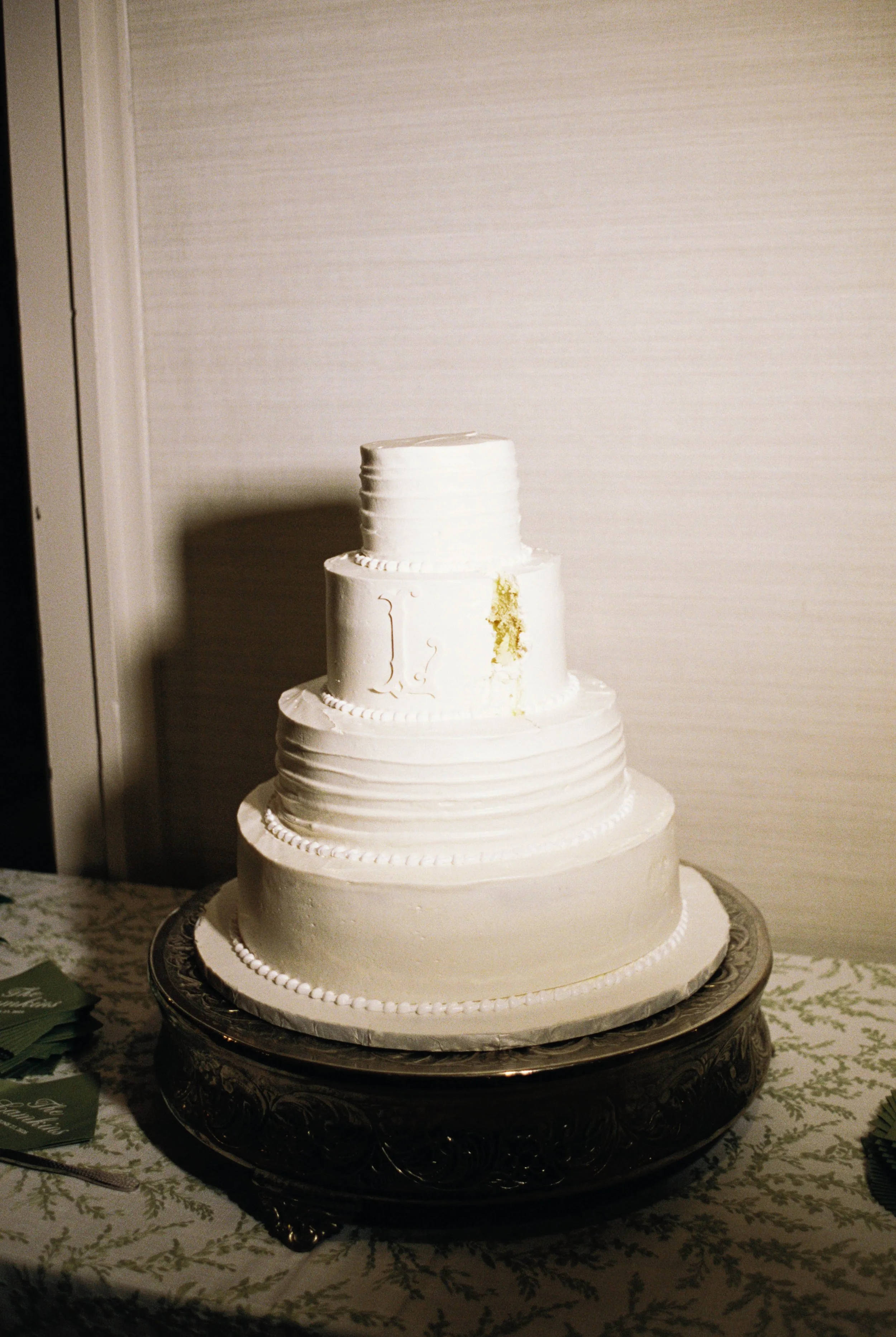 A four-tiered white wedding cake with decorative patterns, placed on a silver tray on a table with a patterned tablecloth.