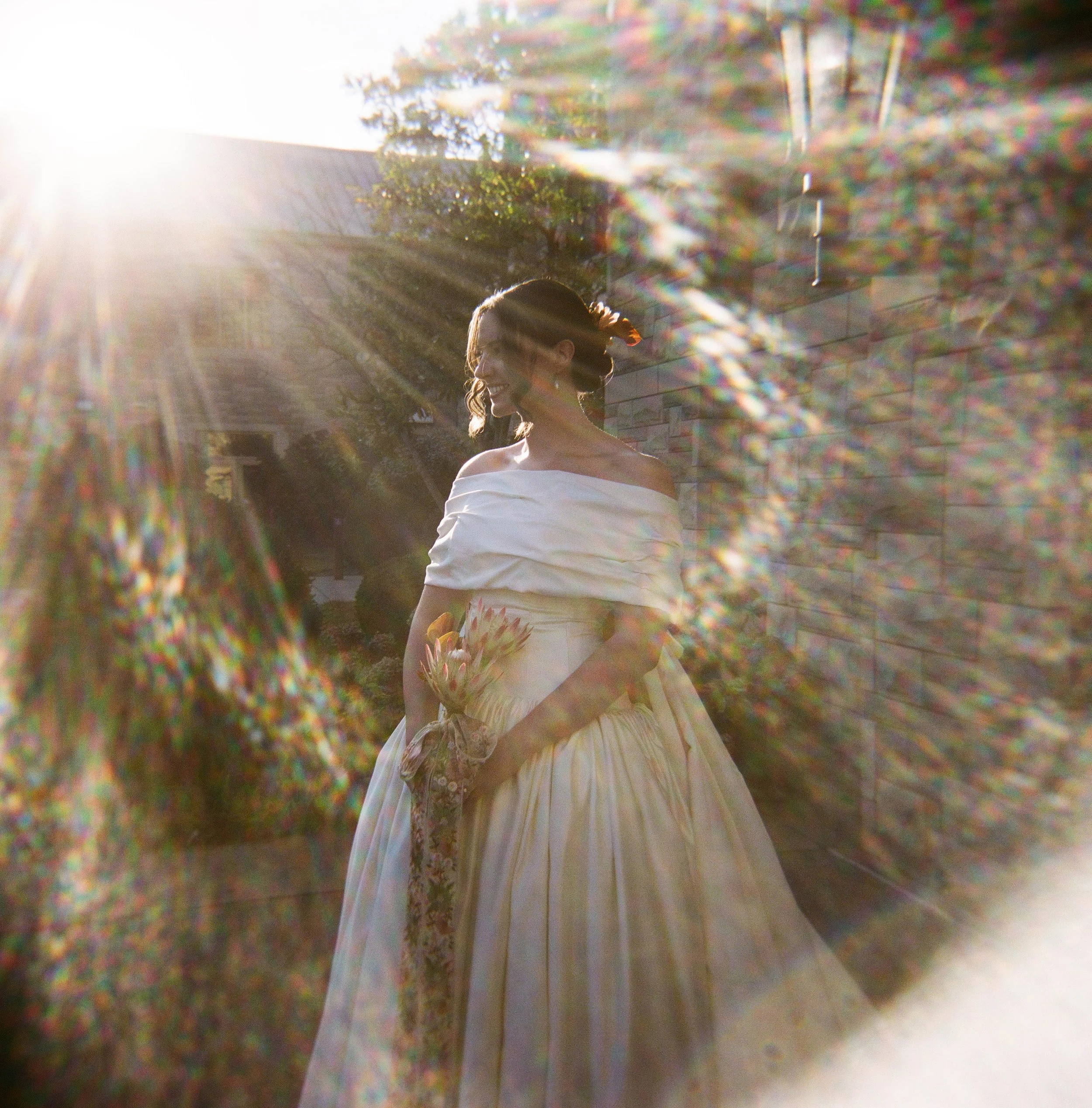 A woman in a white off-the-shoulder dress holding a bouquet of flowers, standing outside near a brick wall with sunlight shining behind her.