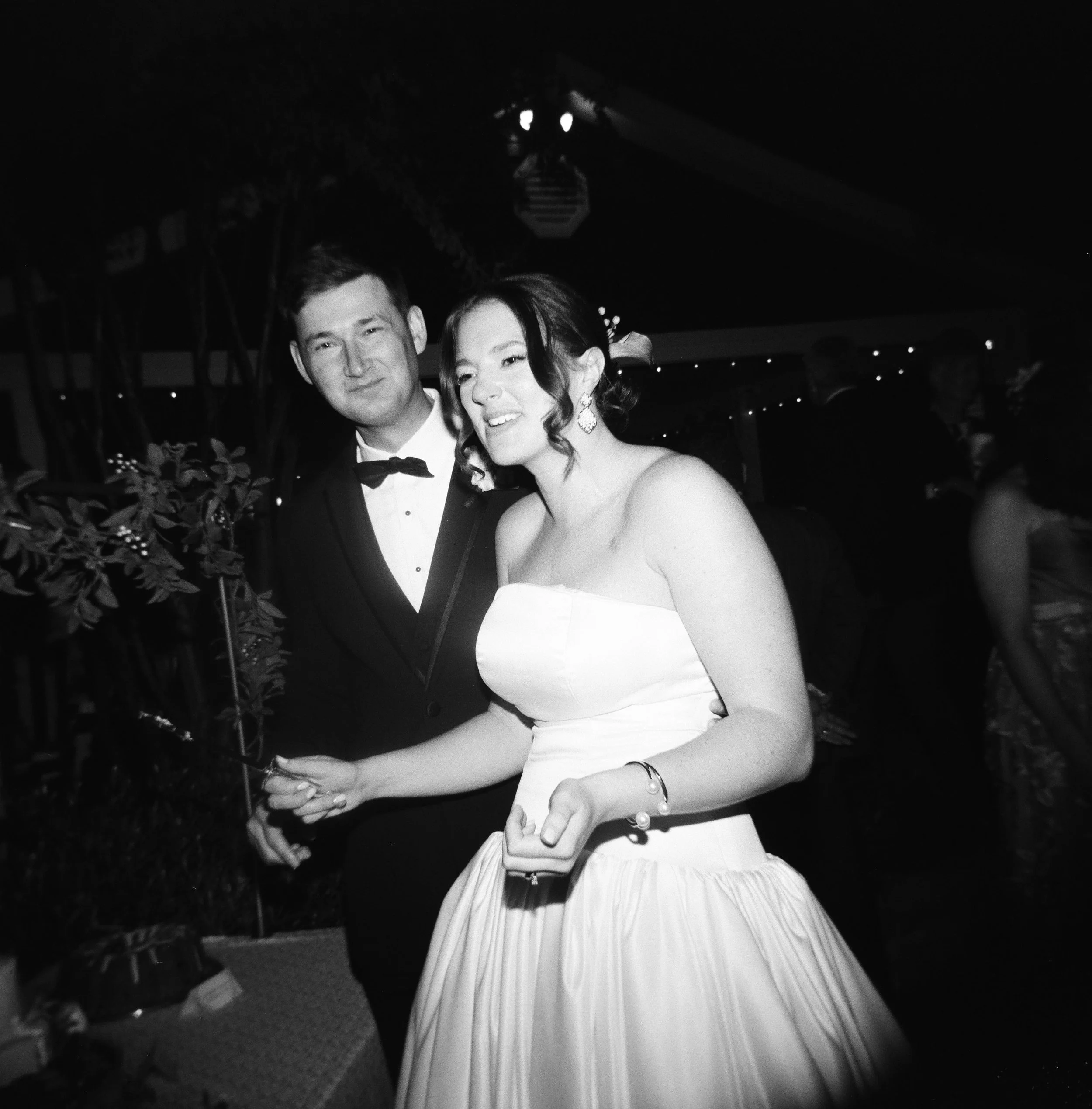 A black and white photo of a man and woman at a formal event. The man is wearing a tuxedo with a bowtie, and the woman is in a strapless gown with earrings and a bracelet. They are smiling and holding hands.