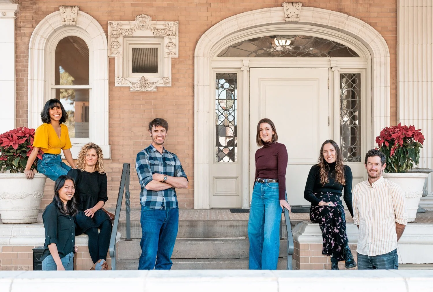 Group of seven diverse young adults standing and sitting on the steps in front of a brick building with large windows and decorative architectural elements, smiling for the photo.
