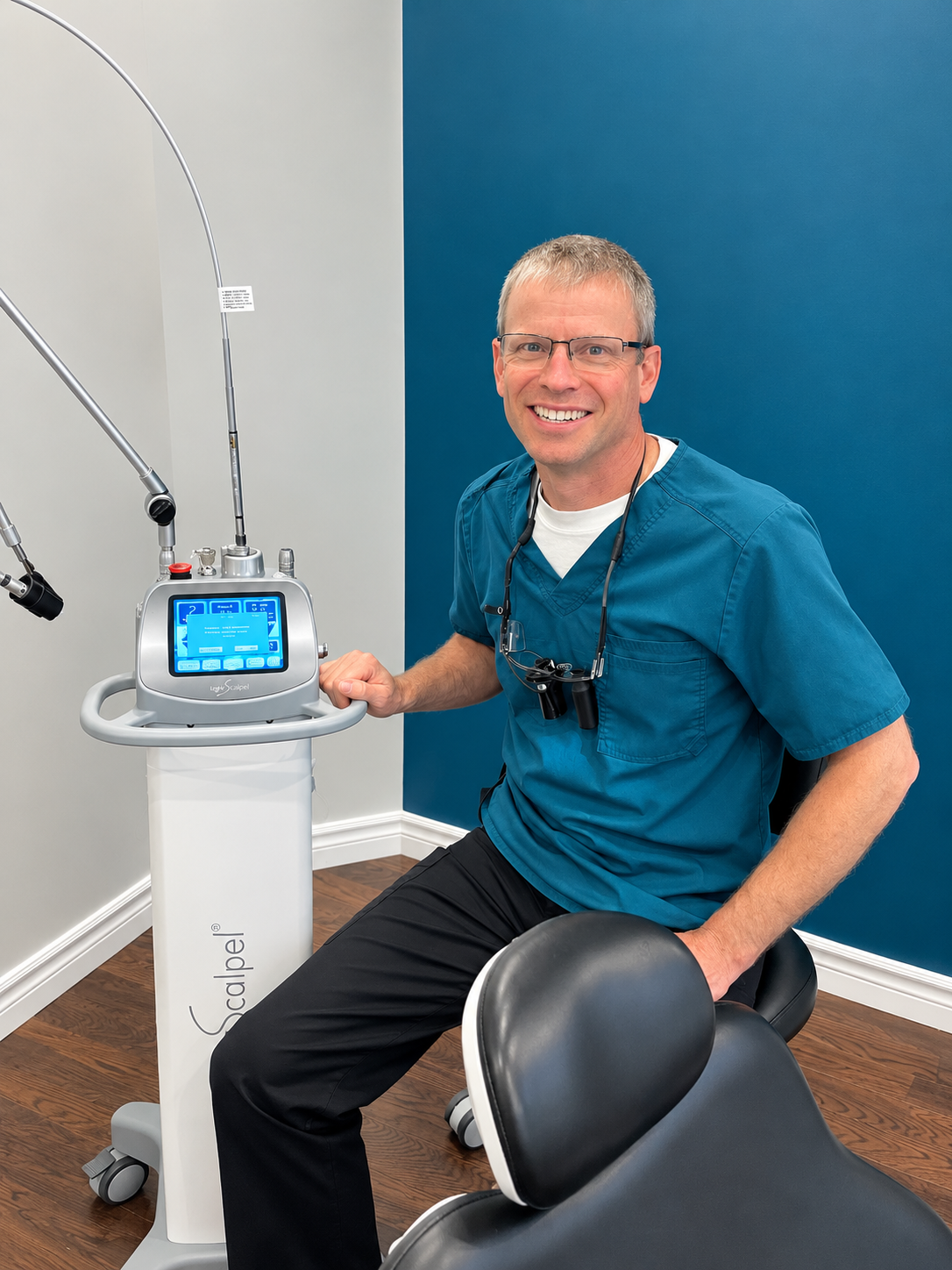 A smiling male healthcare professional in blue scrubs wearing glasses and a stethoscope around his neck, sitting next to medical equipment in a clinical setting with a blue accent wall behind him.