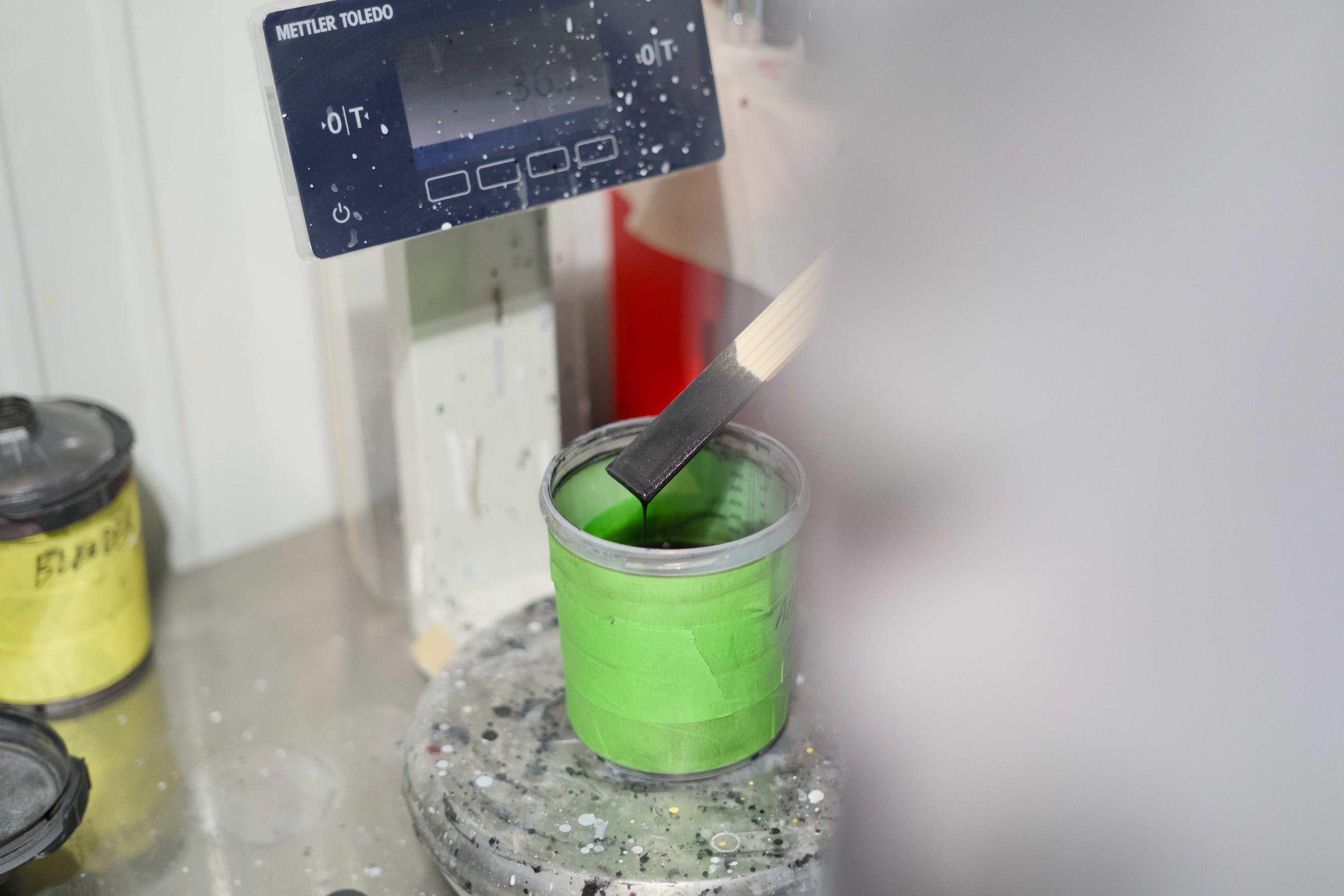 A small container with green paint, with a paintbrush dipped into it, set on a speckled table surface. Above, a digital device is attached to a wall, displaying measurements, with water droplets on its surface.