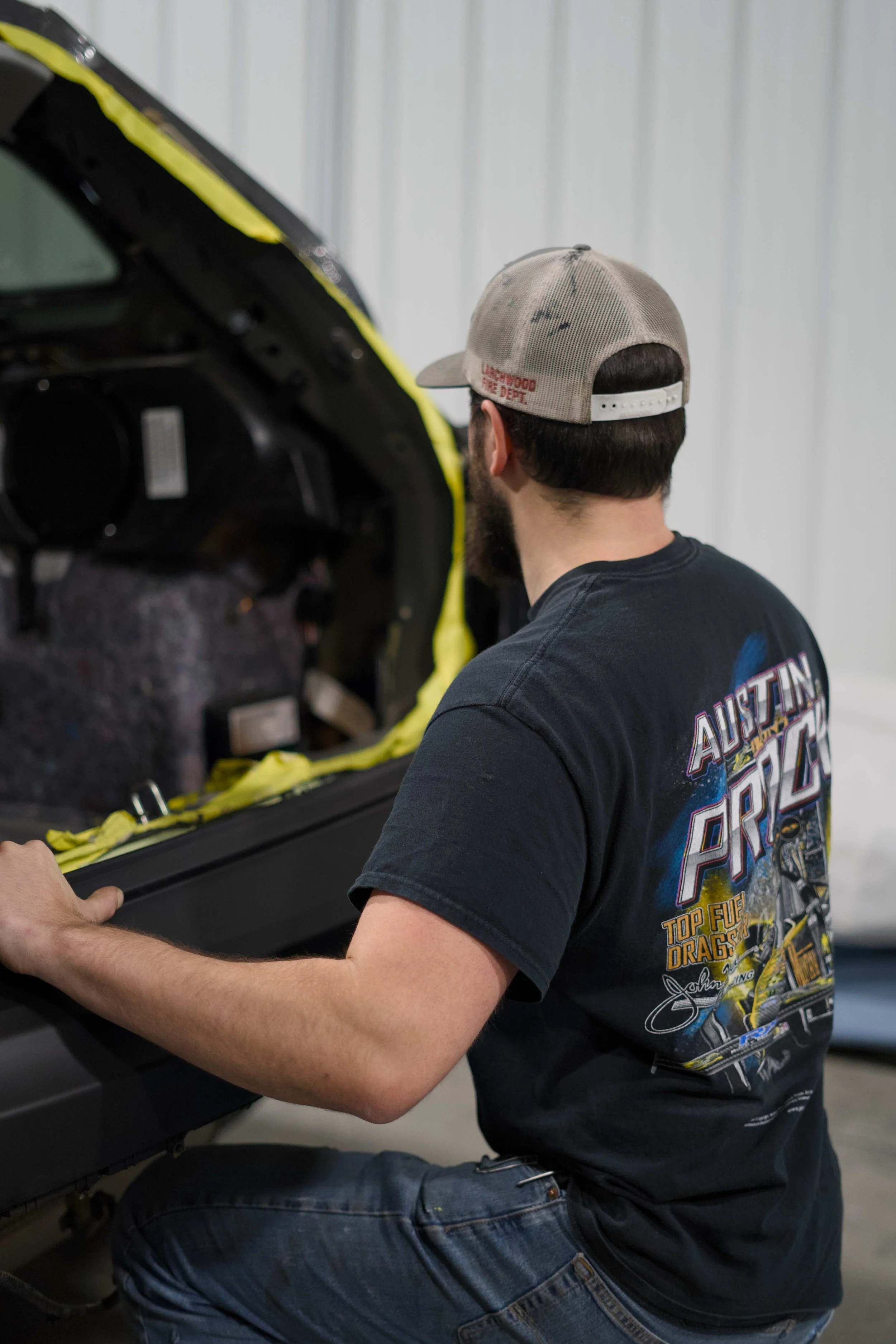 A man with a beard wearing a black Austin Powers T-shirt, blue jeans, and a beige baseball cap working on a car engine in a garage.