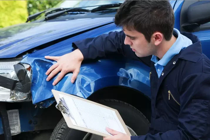 A man inspecting the front of a blue car with a damaged fender after an accident, holding a clipboard.