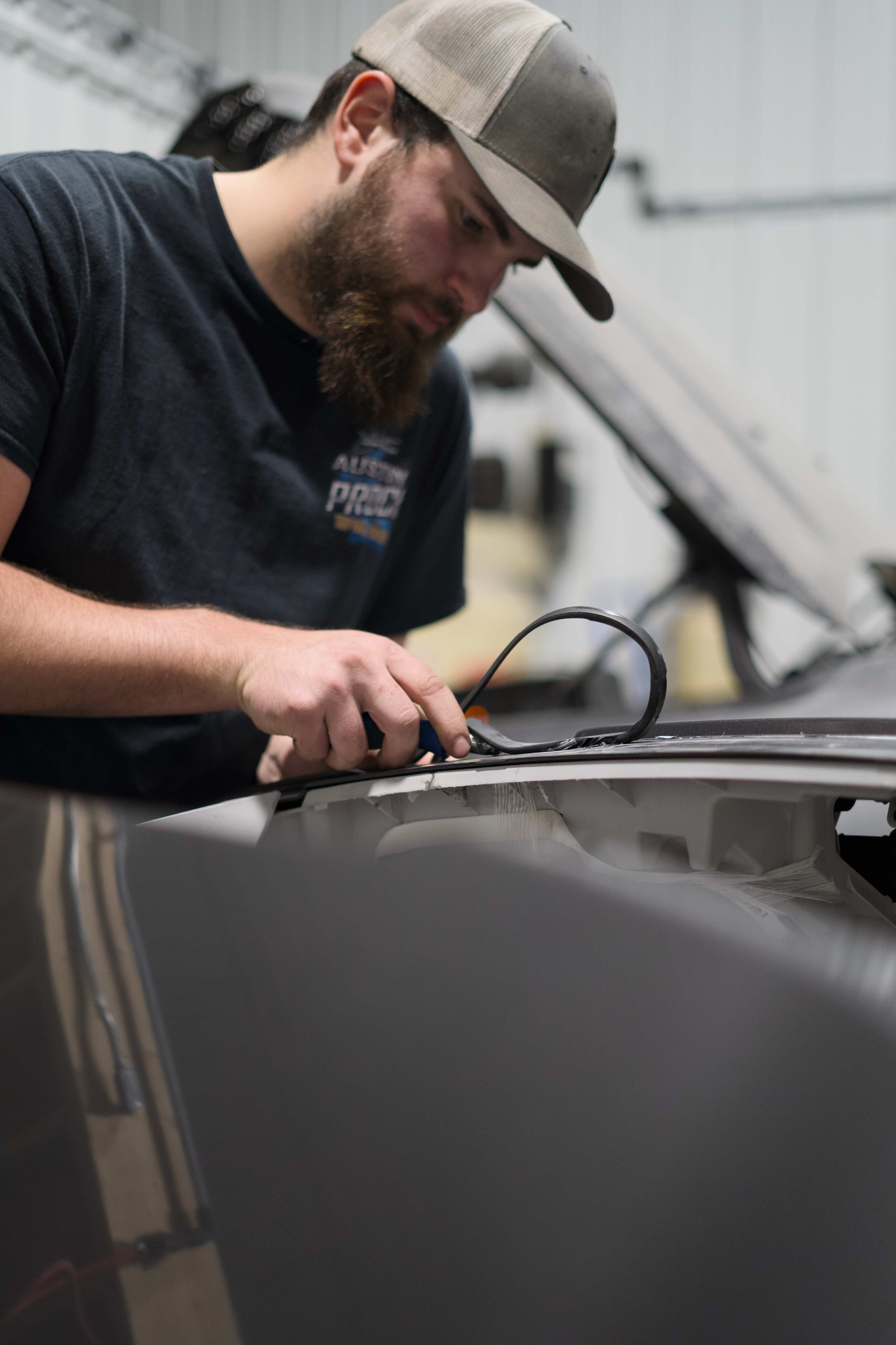 A man with a beard and wearing a beige cap is working on a car inside a workshop. He is using a tool on the car's surface.