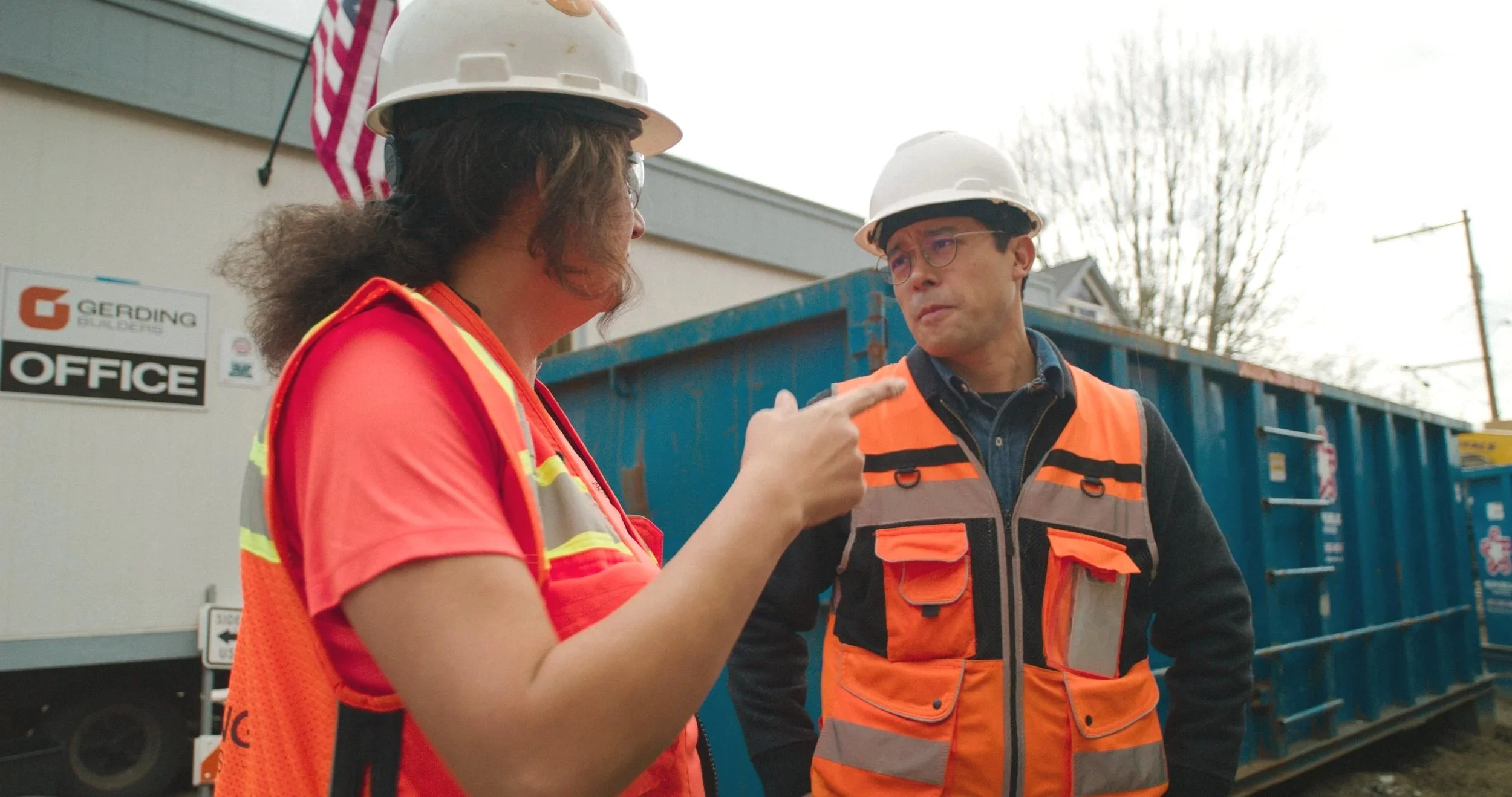 Two construction workers, a woman and a man, are having a conversation outdoors next to a blue construction dumpster. The woman is pointing at the man, who is listening attentively. Both are wearing safety helmets and orange safety vests.