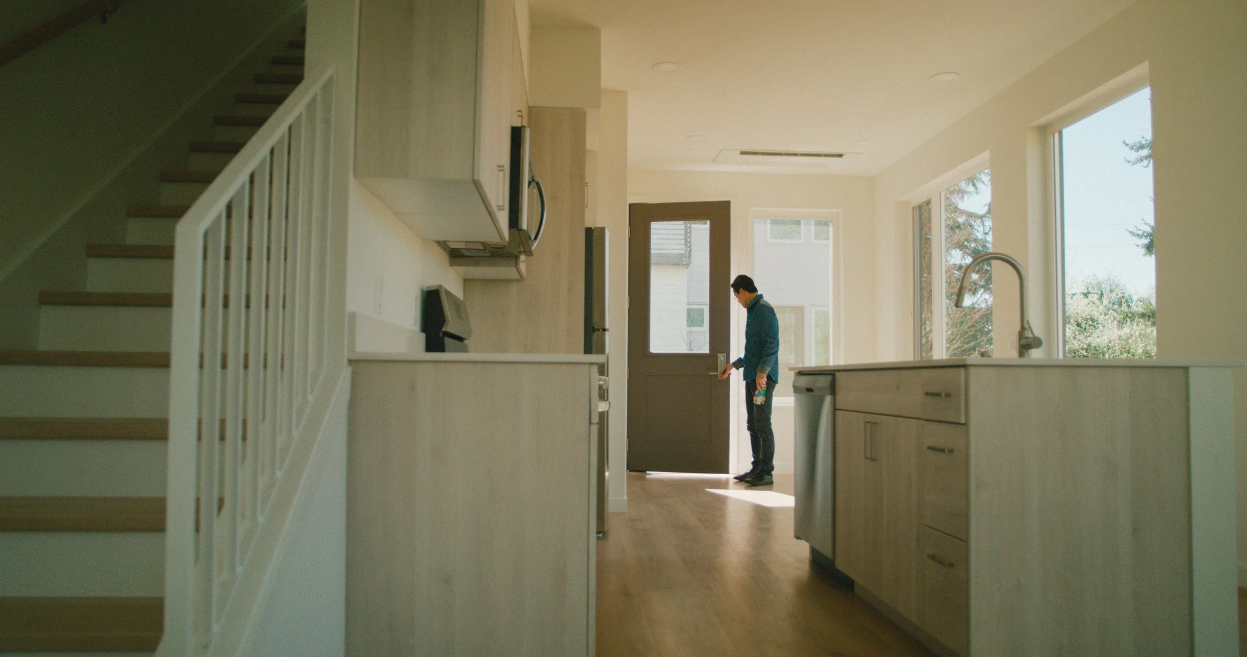 Interior view of a modern kitchen with a person standing near the door, holding a cleaning spray, surrounded by large windows and white cabinets.