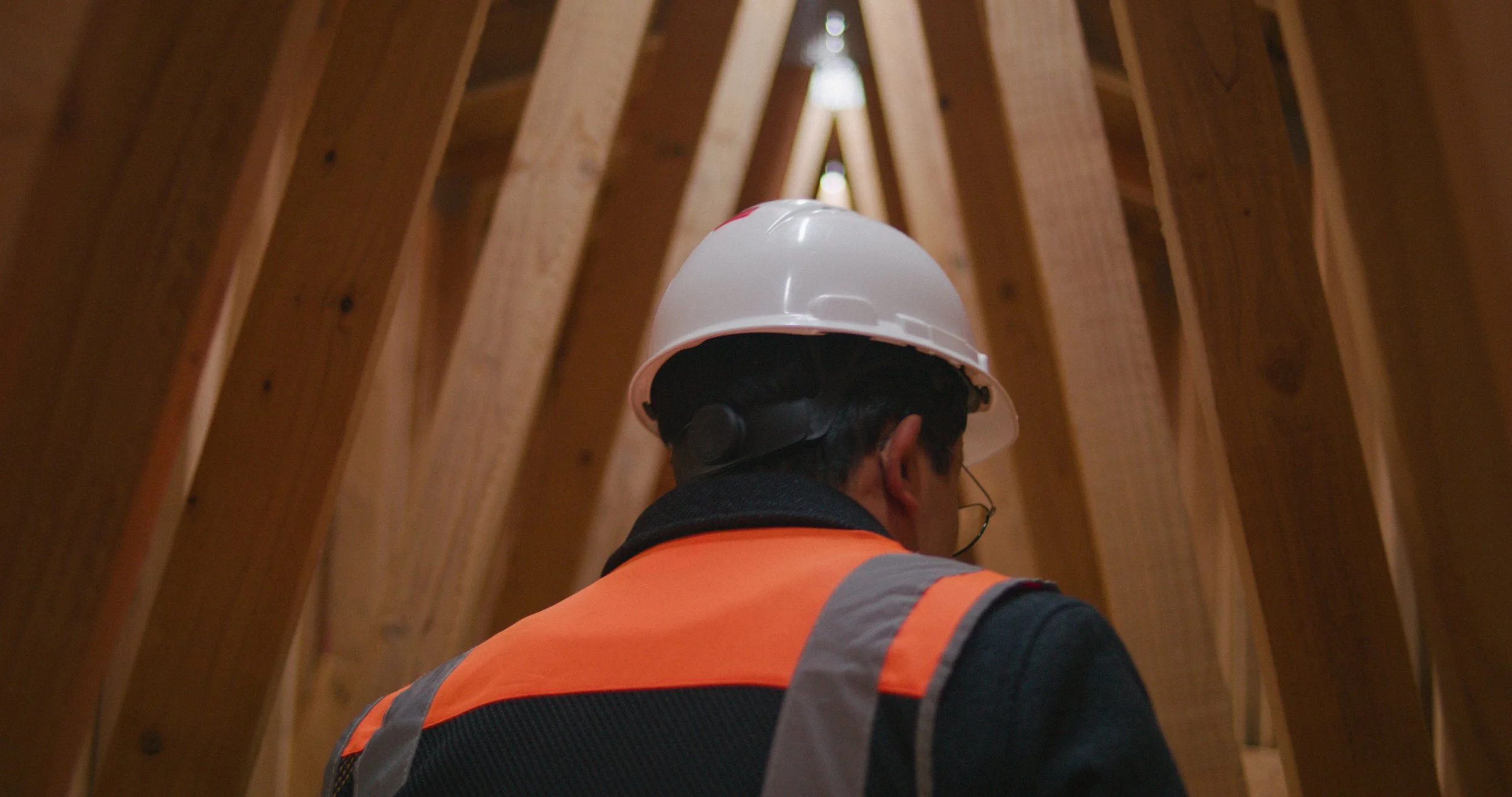 A construction worker wearing a white safety helmet and an orange safety vest inside a wooden frame structure.