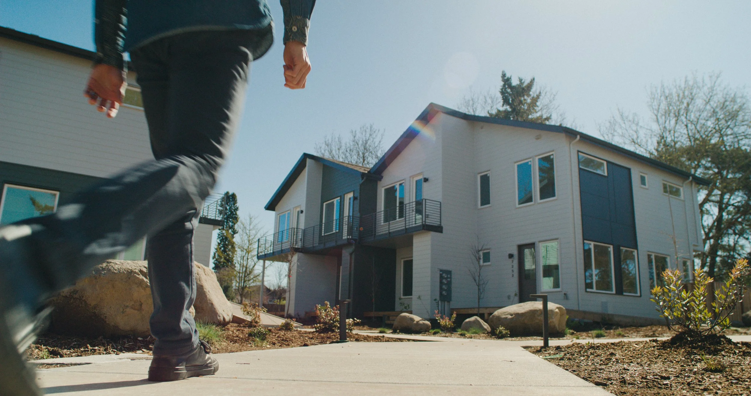 Person walking on a sidewalk in front of a modern, multi-story house with large windows and a balcony, under a clear sky with trees in the background.