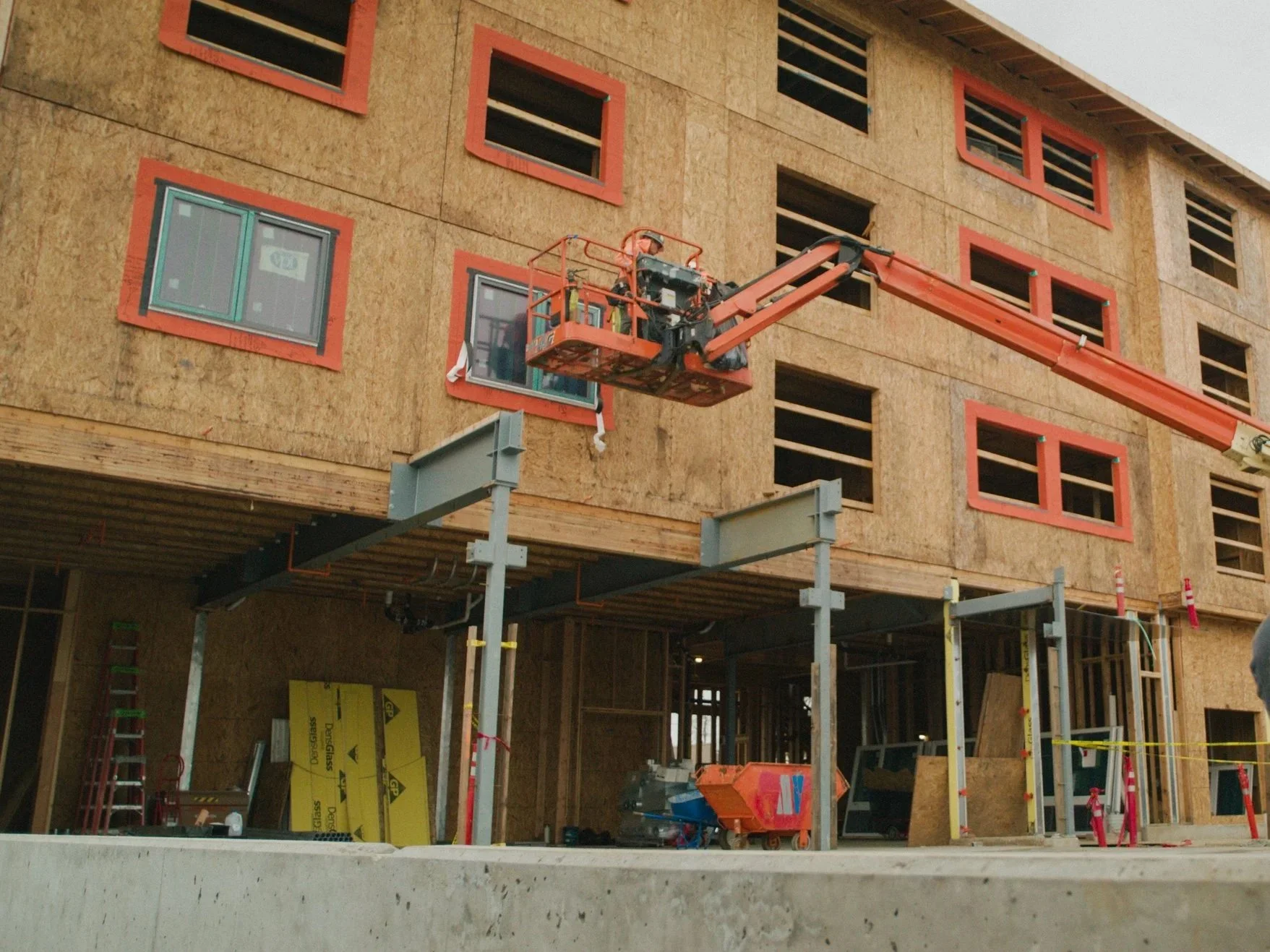 Construction site with a building under construction, orange-framed windows, and workers on a cherry picker lift.