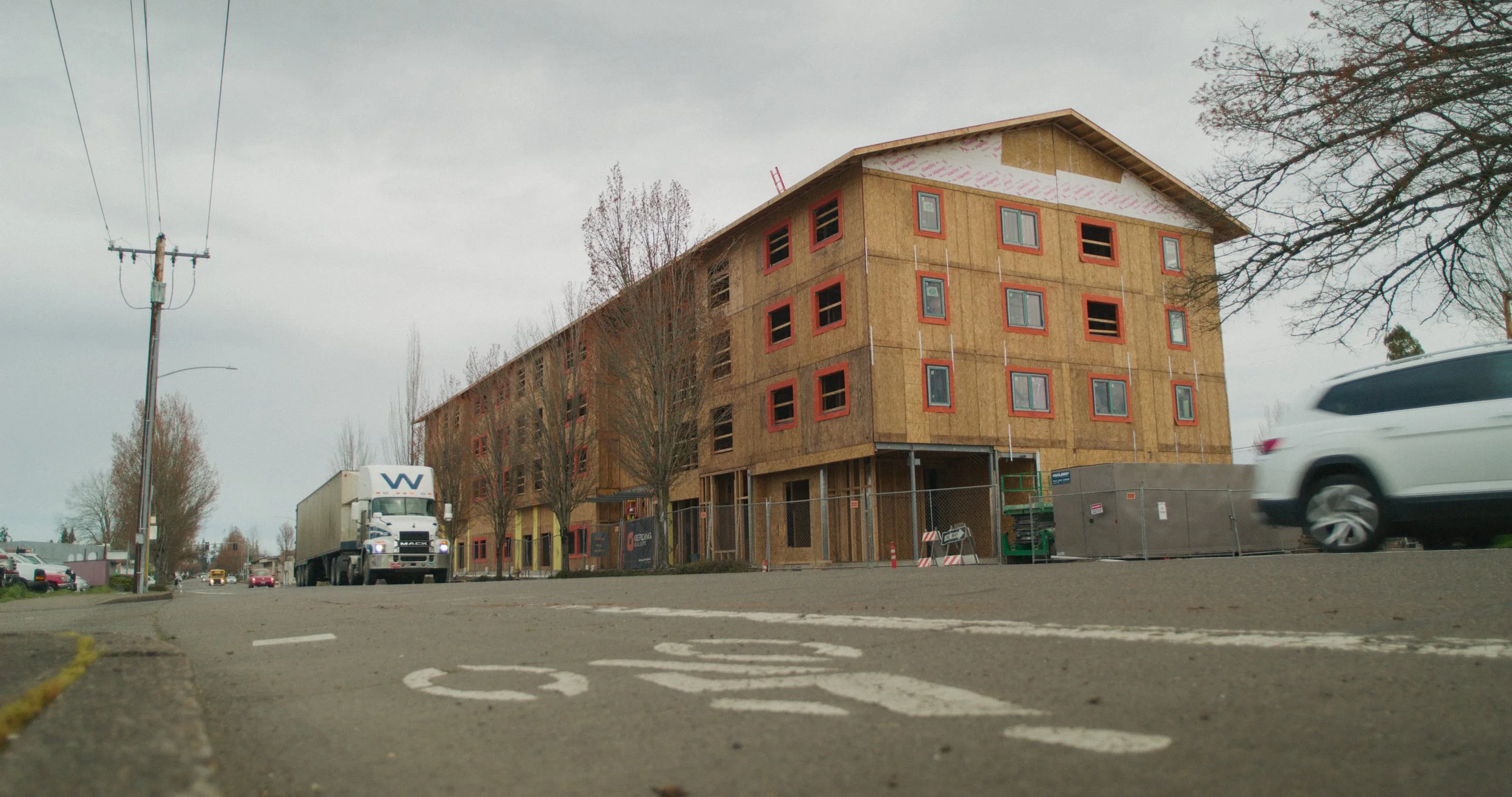A multi-story building under construction with a wooden frame and red window borders, located on a city street with parked cars, trees, and construction fencing.